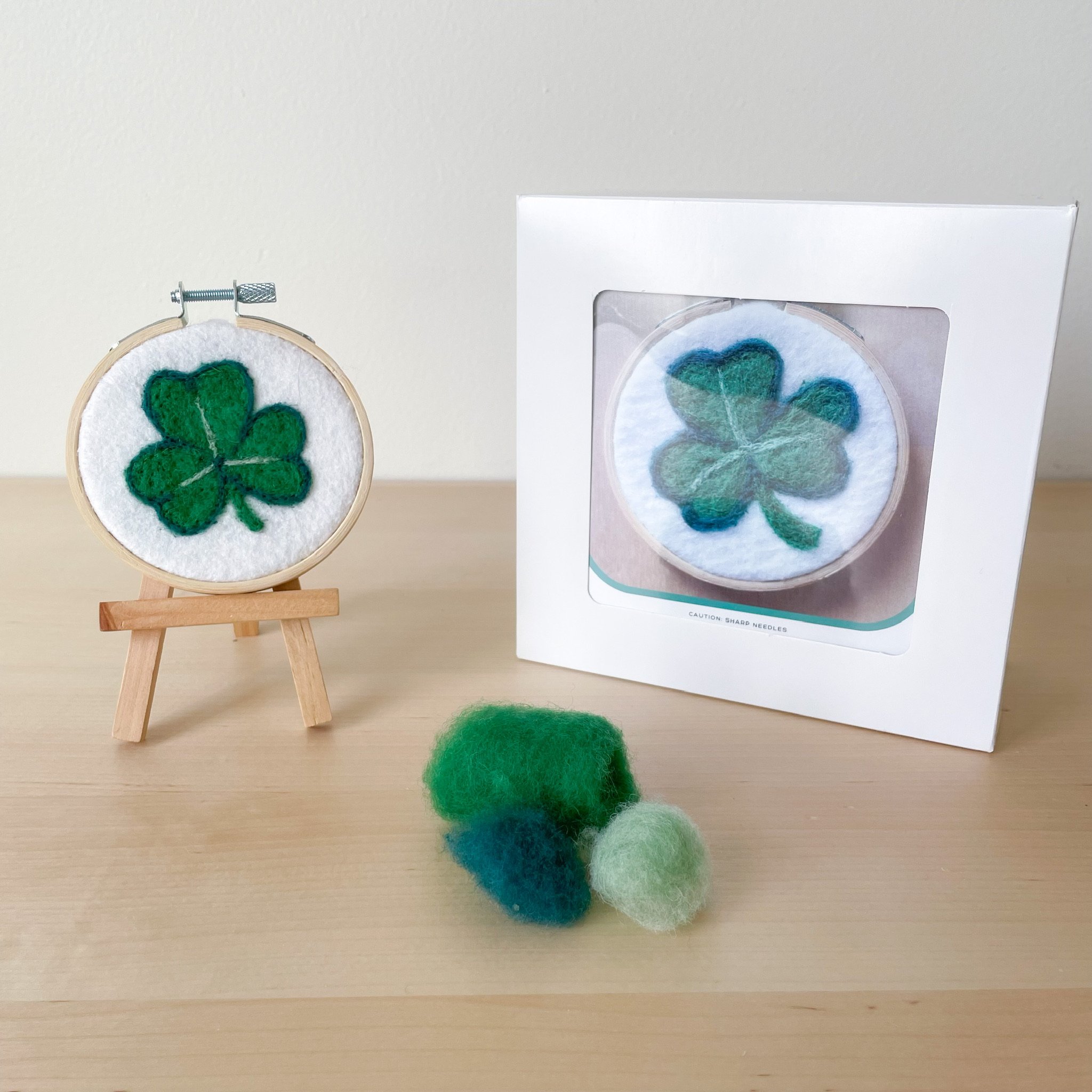 Felted shamrock art framed in an embroidery hoop sitting on a mini easel, in front of the white box of the kit and tufts of green wool.