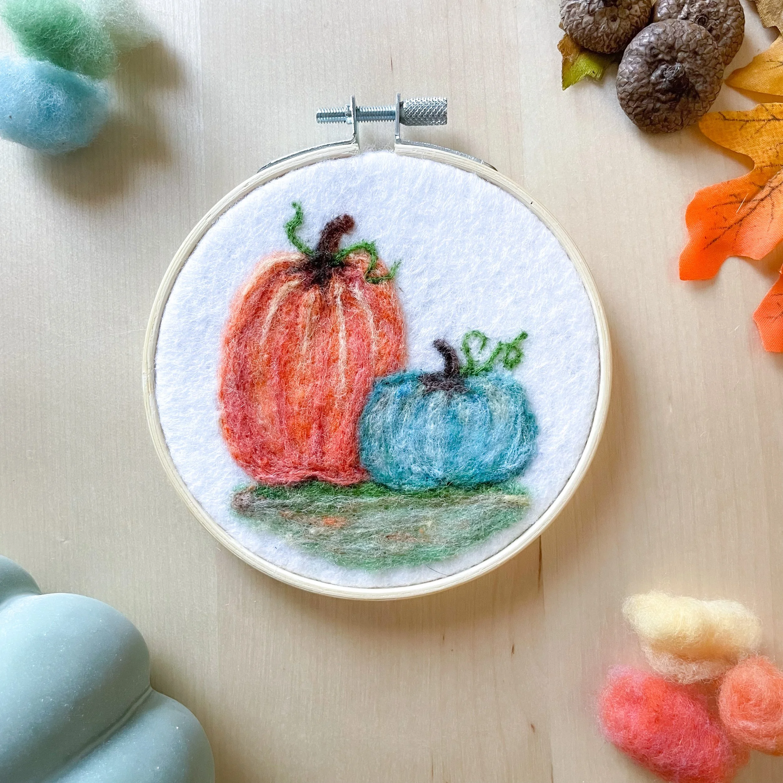 Felted artwork of an orange and teal pumpkin framed in a wood hoop, laying on a light wood table surrounded by fall leaves, acorns, and tufts of colorful wool.