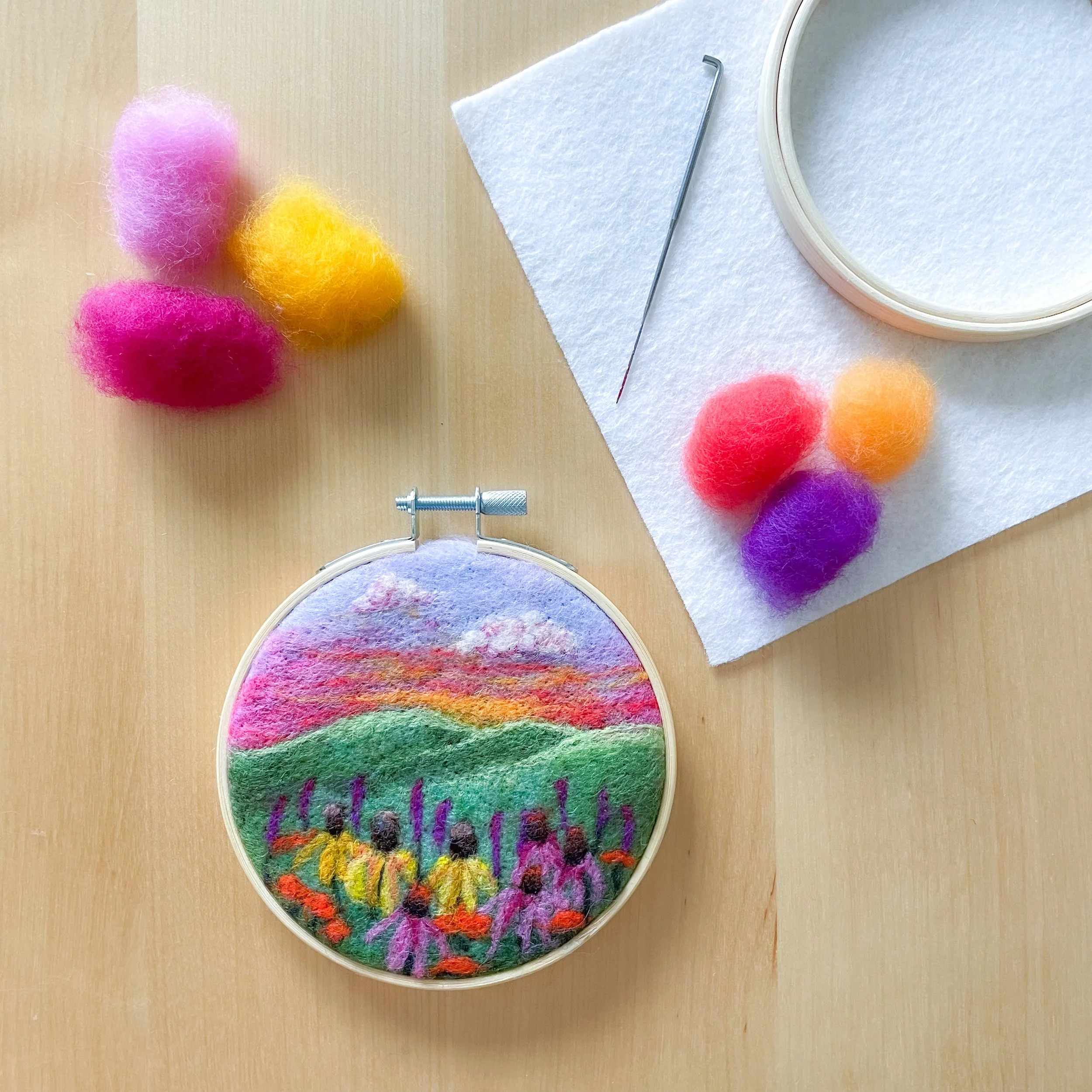 Colorful felted art of a sunset and prairie flowers framed in a hoop, sitting on a wood table with colorful tufts of wool, white felt, a hoop and needle surrounding it.