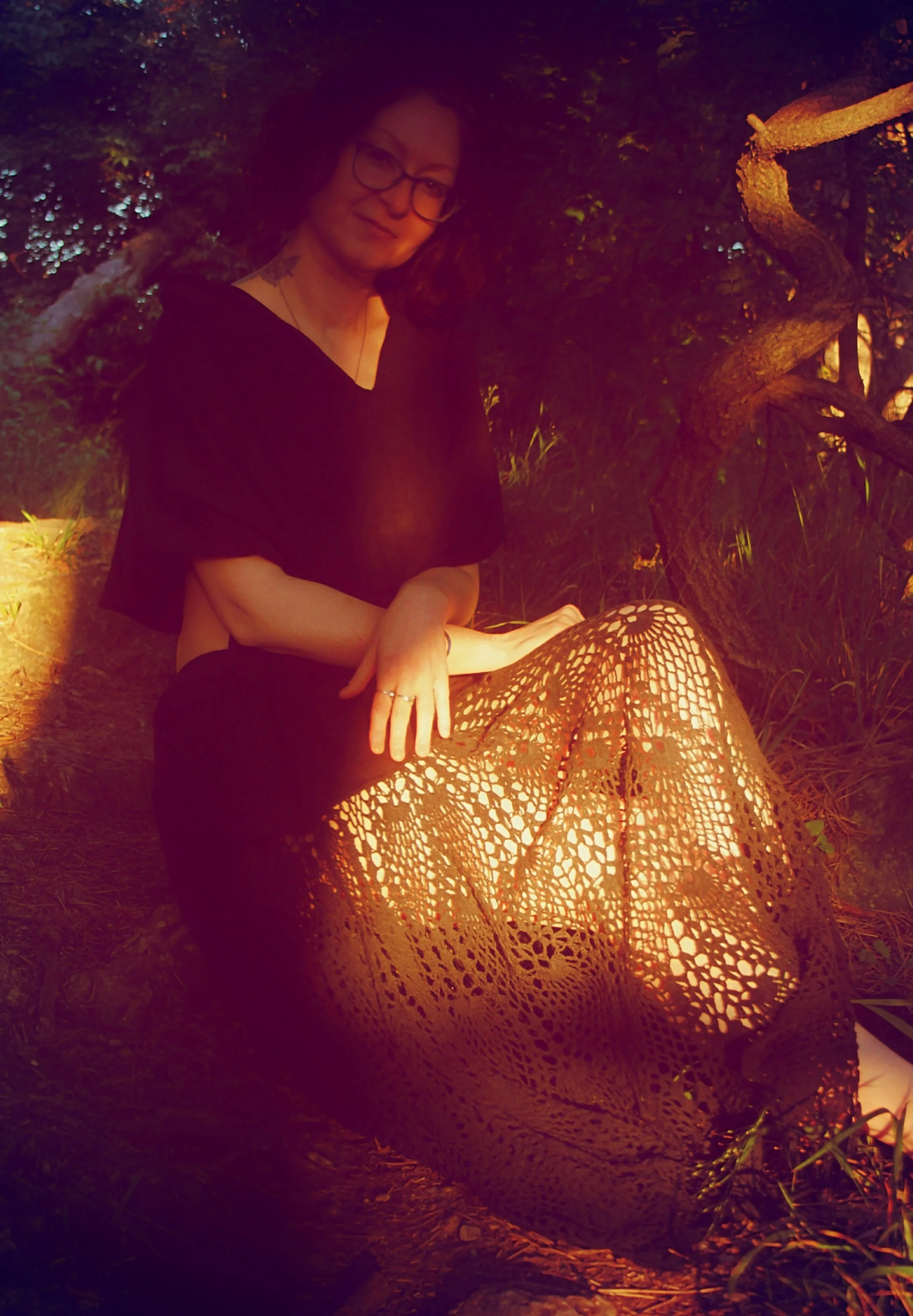 A woman sitting outdoors at dusk or night, wearing glasses, a dark top, and a crochet skirt, with trees and a twisted tree branch behind her.
