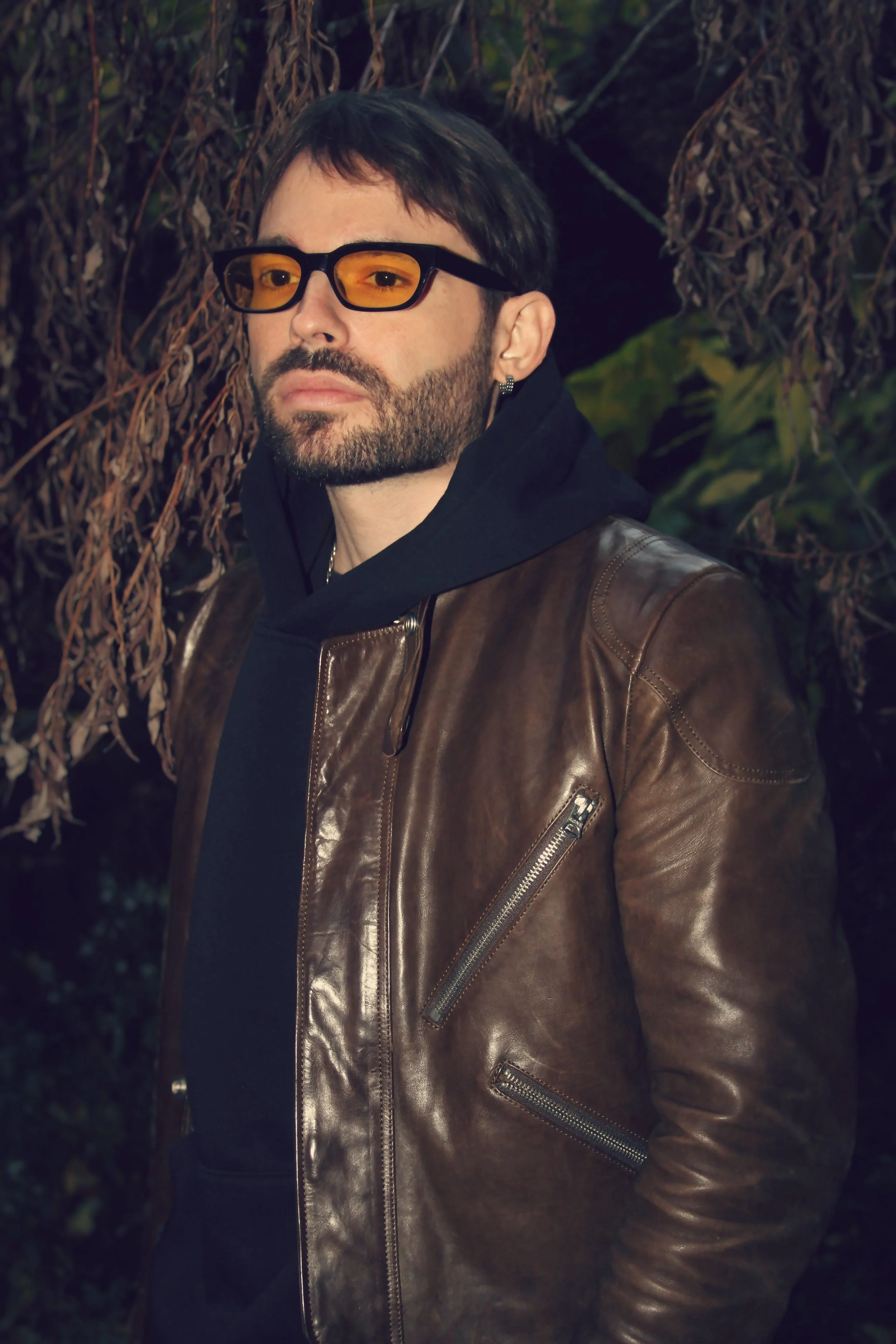 A man with dark hair, beard, and wearing sunglasses, a black hoodie, and a brown leather jacket standing outdoors near trees.