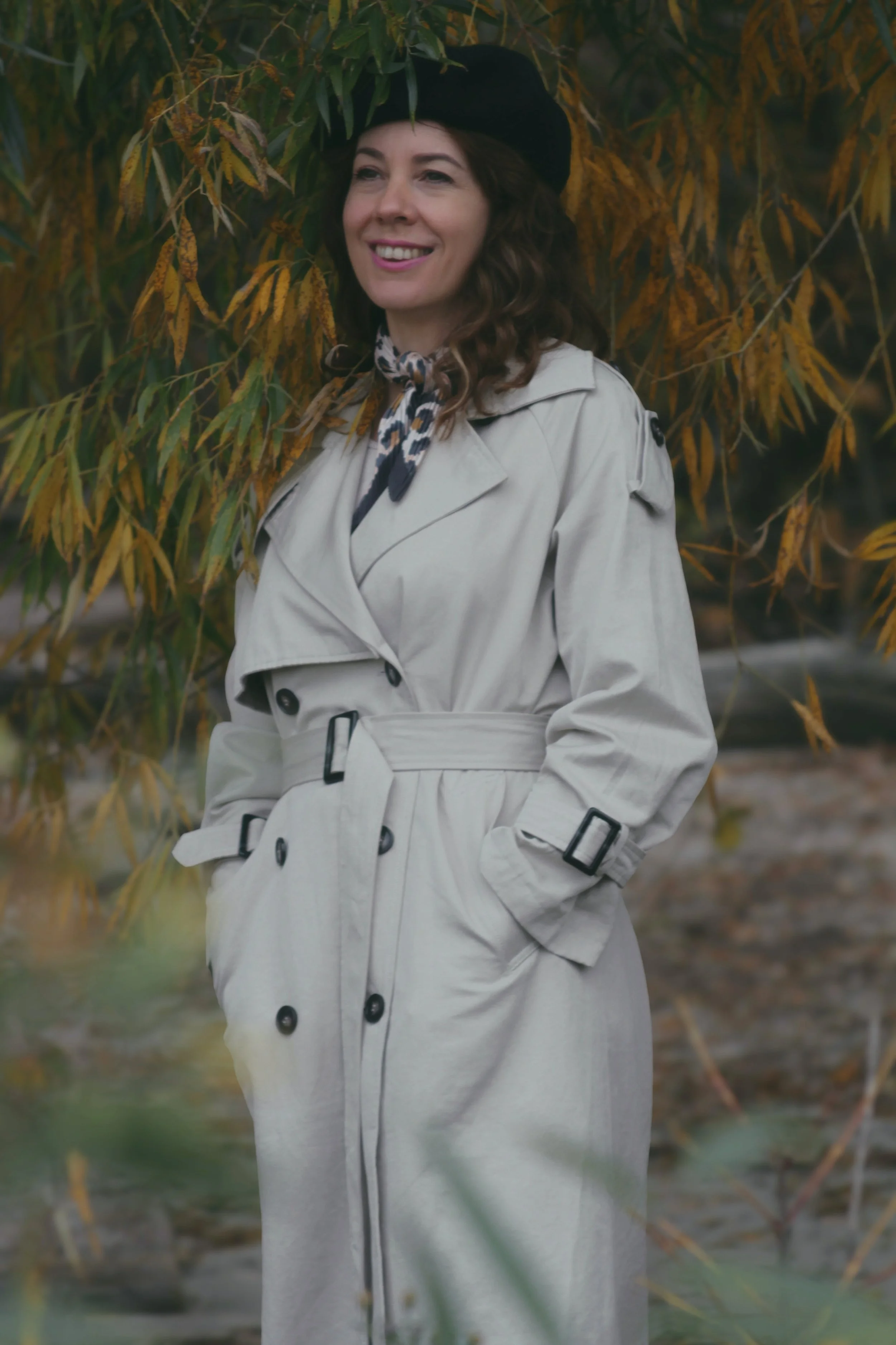 A woman wearing a beige trench coat, a black beret, and a leopard-print scarf, smiling outdoors near autumn foliage.