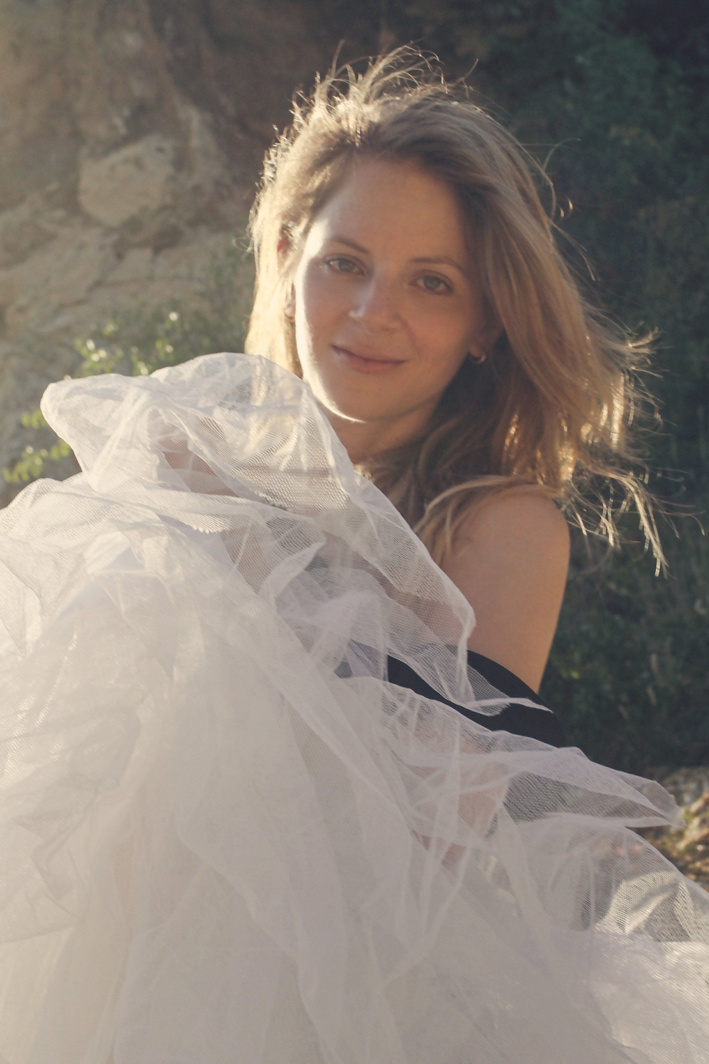 A young woman with shoulder-length hair smiling outdoors with sunlight shining on her face, wearing a dress with white tulle fabric.