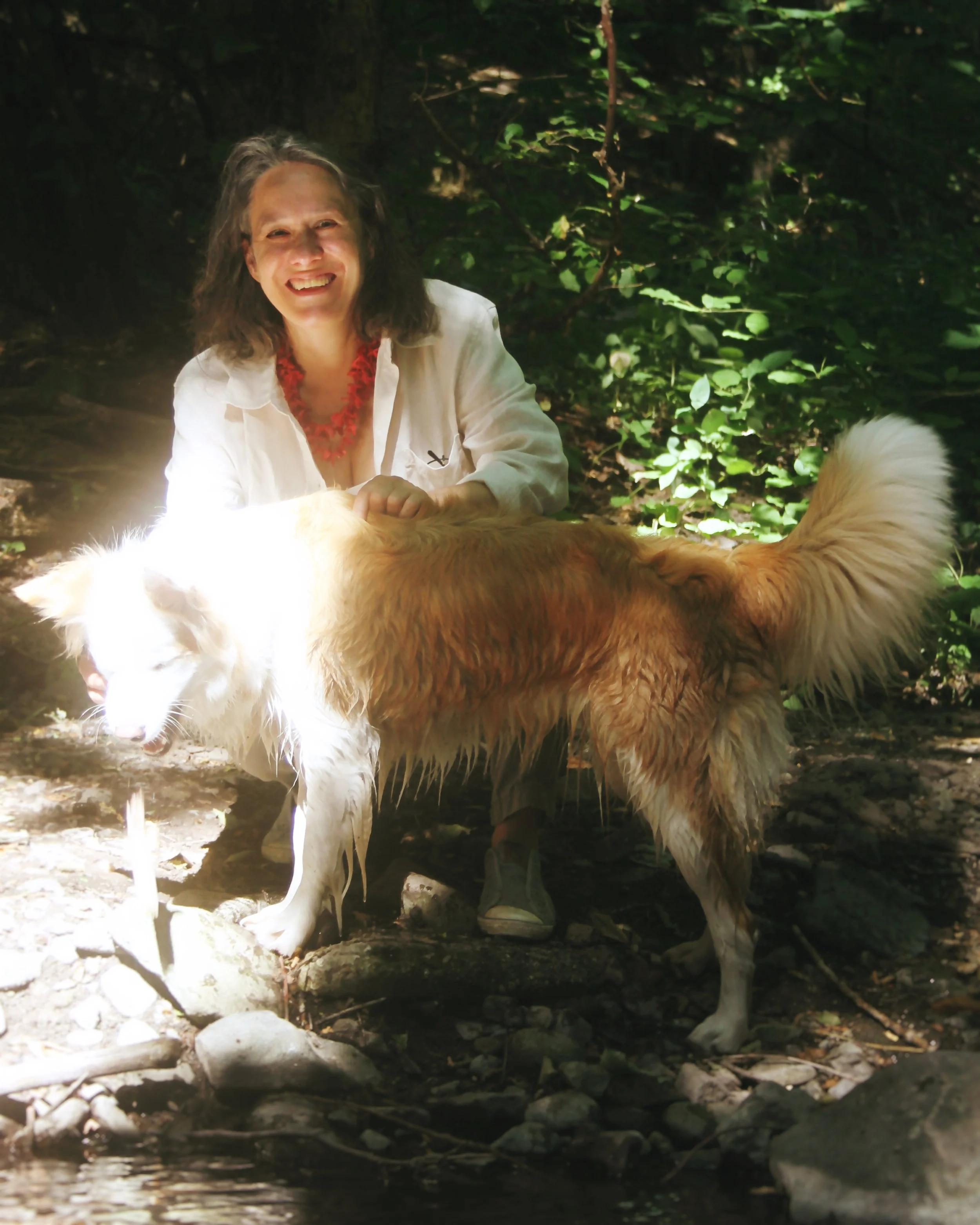 A woman with curly hair wearing a white shirt and red necklace, smiling while standing in a forest, playing with a wet, golden-colored dog near a small stream.