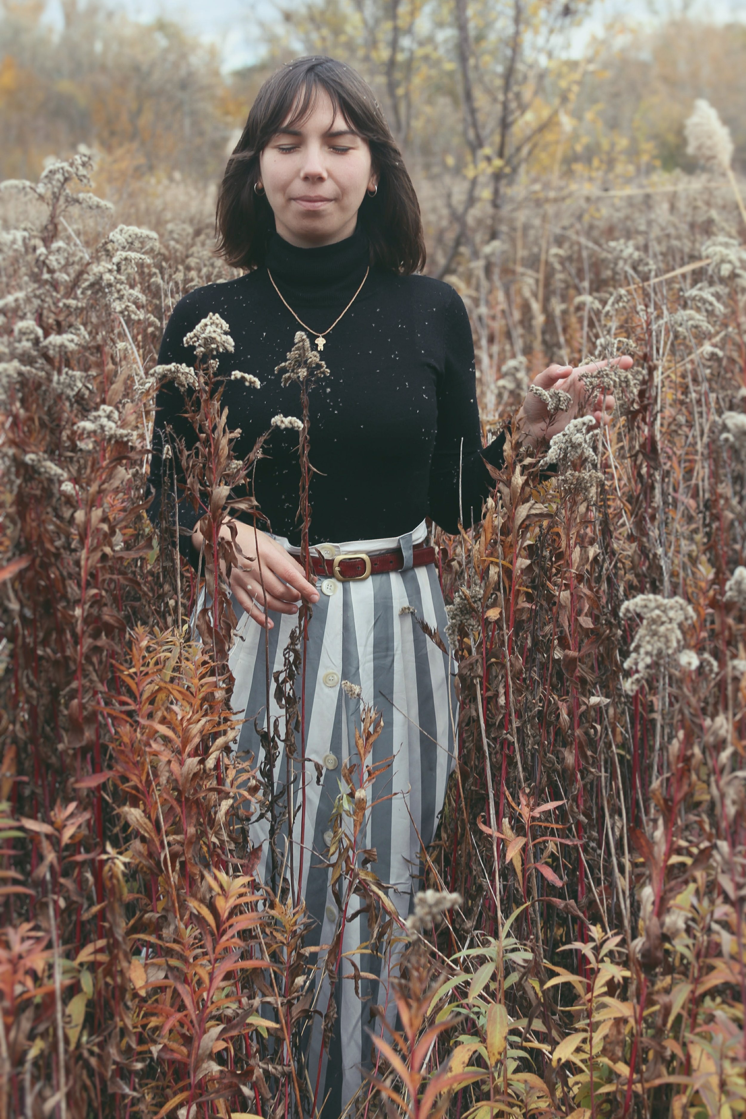 A woman with dark hair and hoop earrings standing in a field of dried plants, wearing a black turtleneck sweater and striped pants with a belt, with trees in the background during autumn.