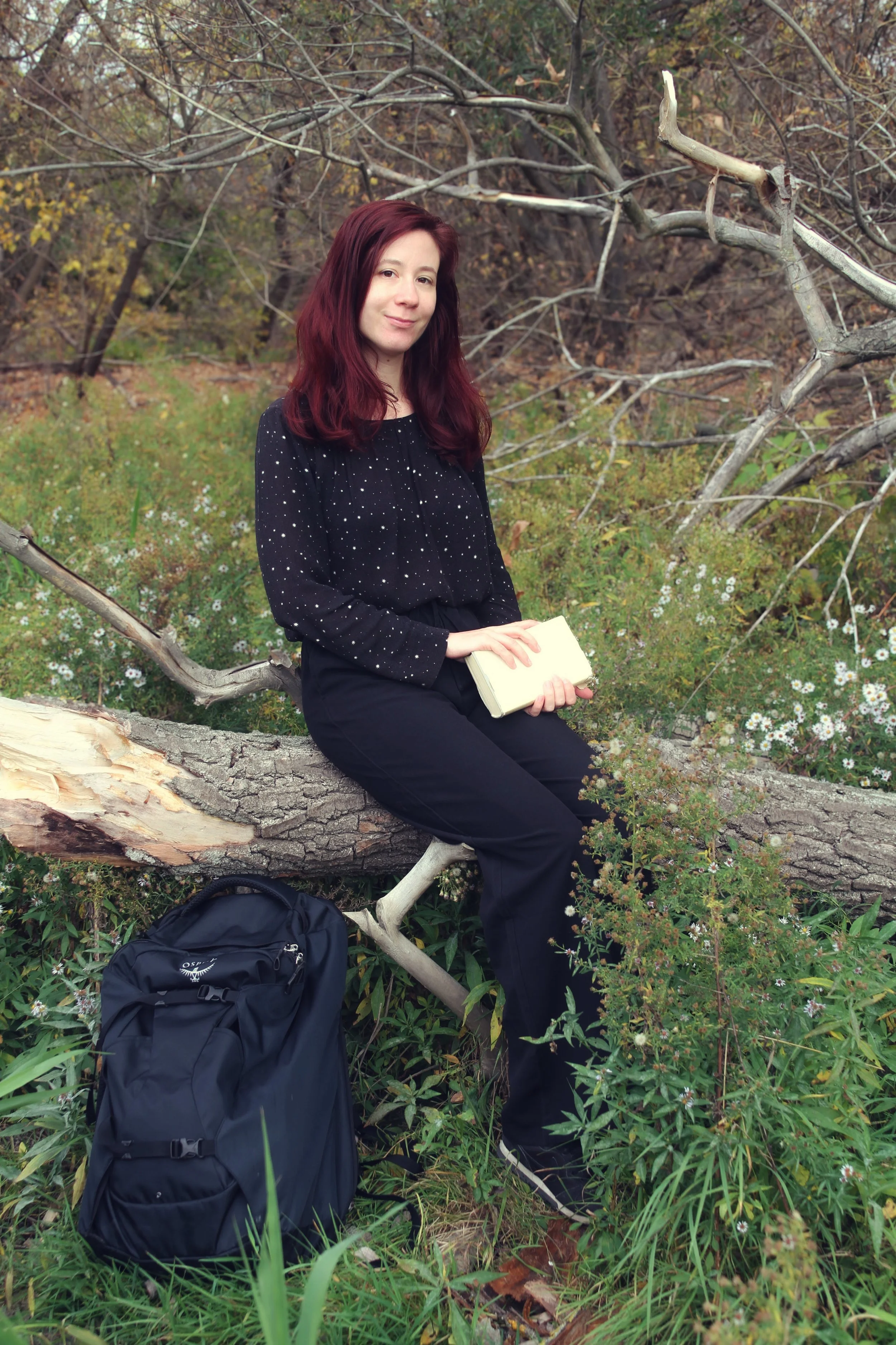 A woman with red hair and black pants sitting on a fallen tree in a forest, holding a closed notebook, with a black backpack on the ground nearby.