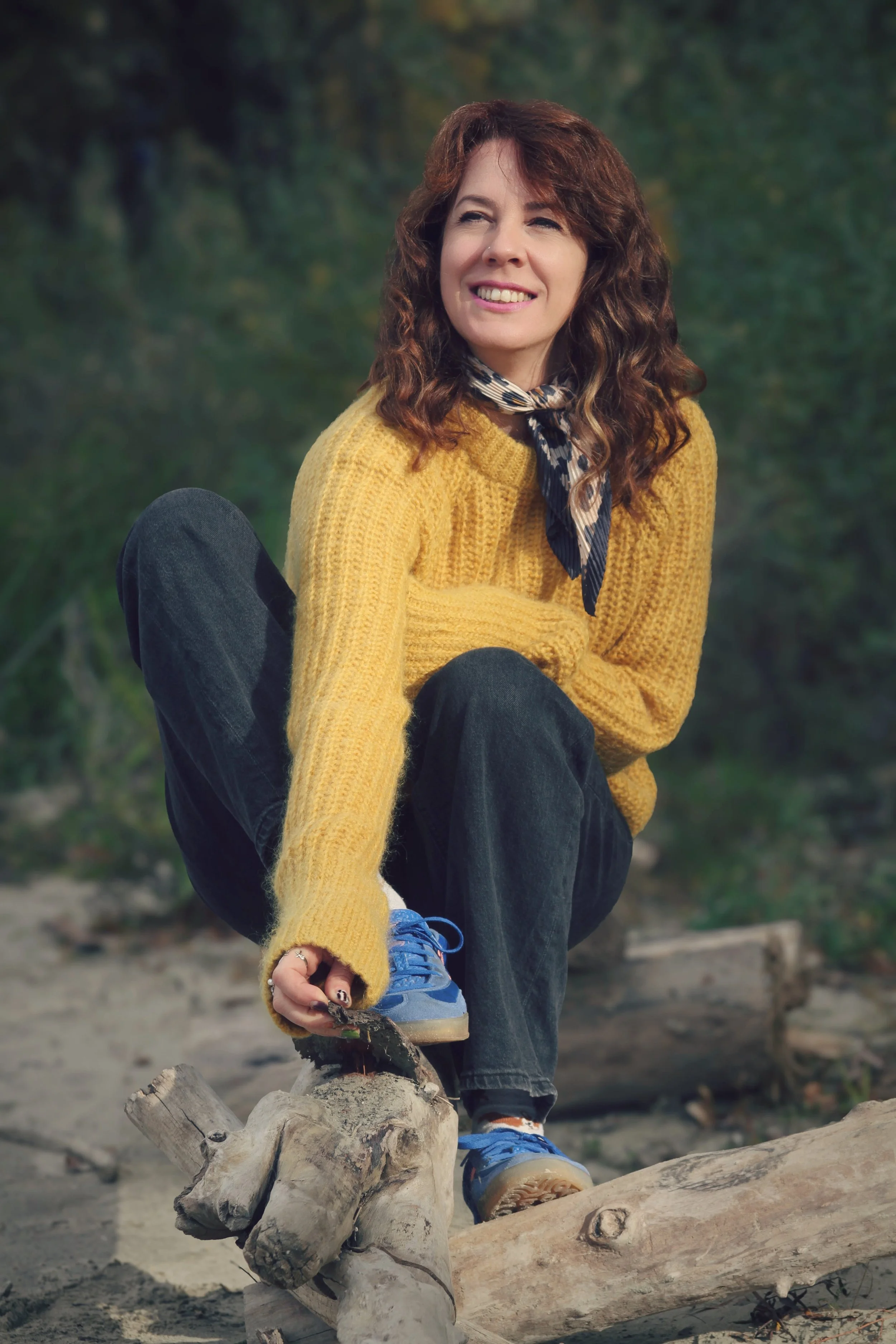 A woman with curly brown hair sitting outdoors on a log, wearing a yellow sweater, a patterned scarf, black pants, and blue sneakers, smiling and adjusting her shoe.