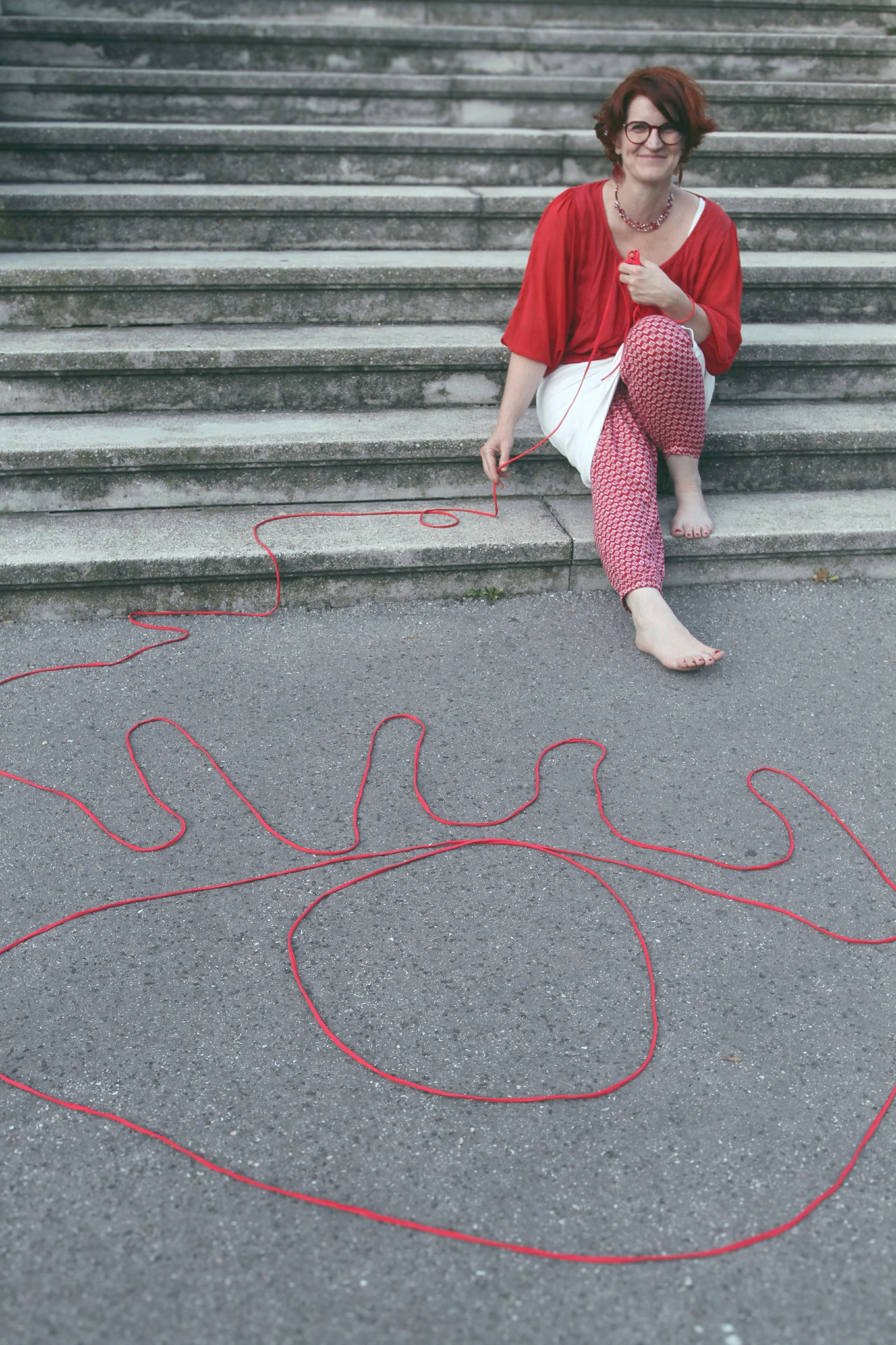 A woman with red hair, glasses, and a red top sitting on outdoor concrete stairs. She is barefoot, holding a piece of red string connected to a spiral shape on the ground, representing a stylized flower or sun.