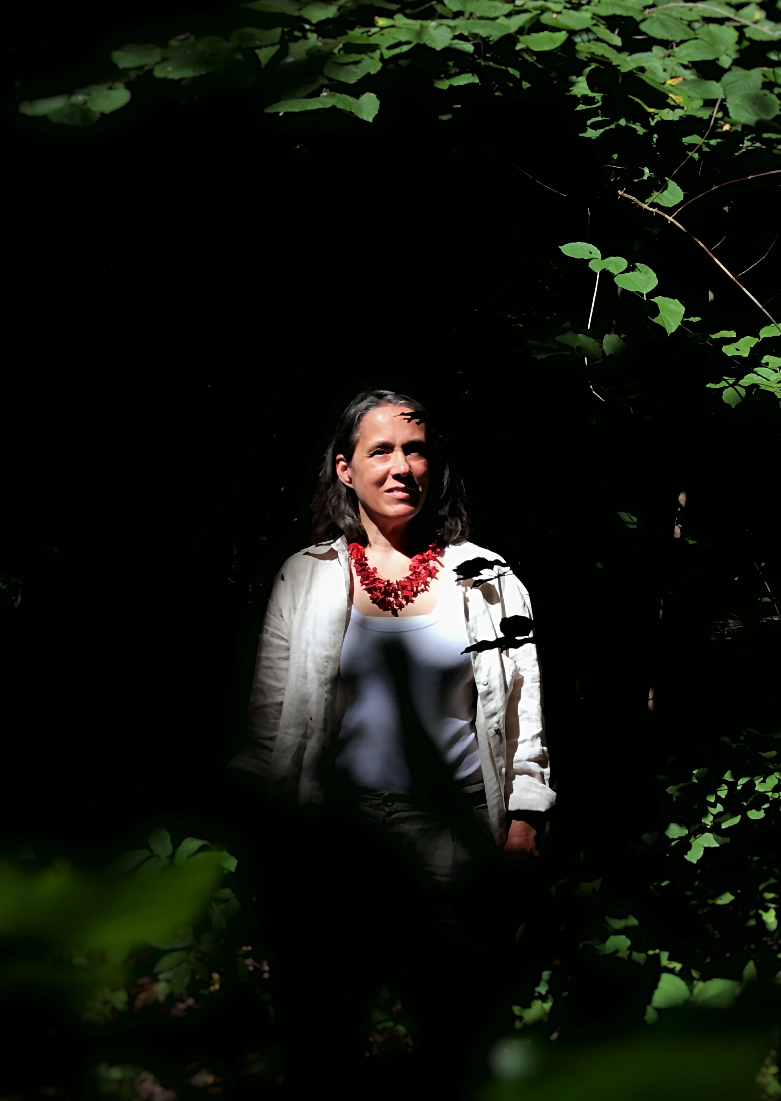 A woman standing in a dark forest or jungle, illuminated by sunlight filtering through the leaves above, wearing a white top, light-colored jacket, and a red necklace.