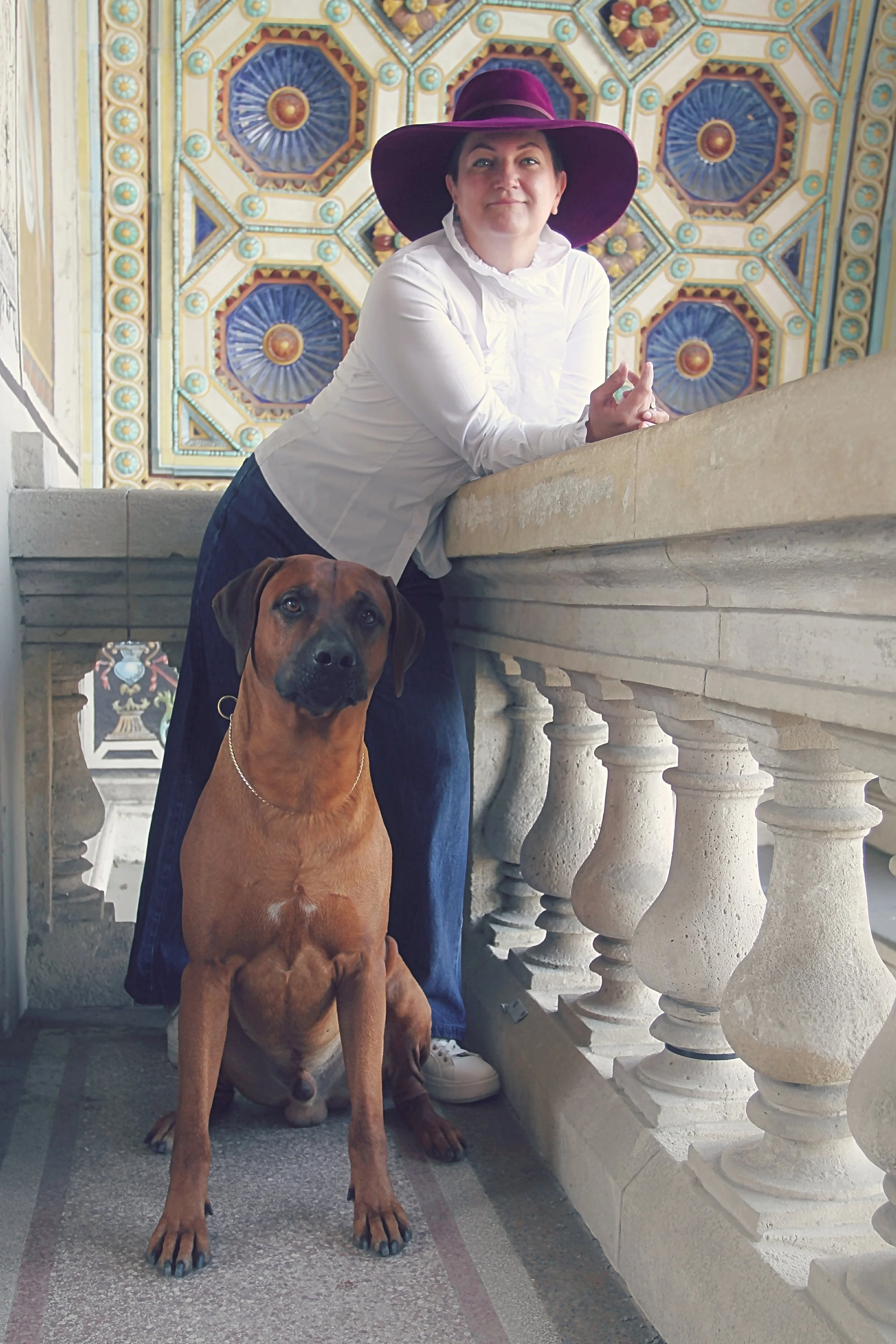 A woman with short dark hair, wearing a white shirt and a large purple hat, leaning on a stone railing inside a building with ornate, colorful, geometric ceiling designs. A brown dog with a black face is sitting on the floor in front of her, facing the camera.