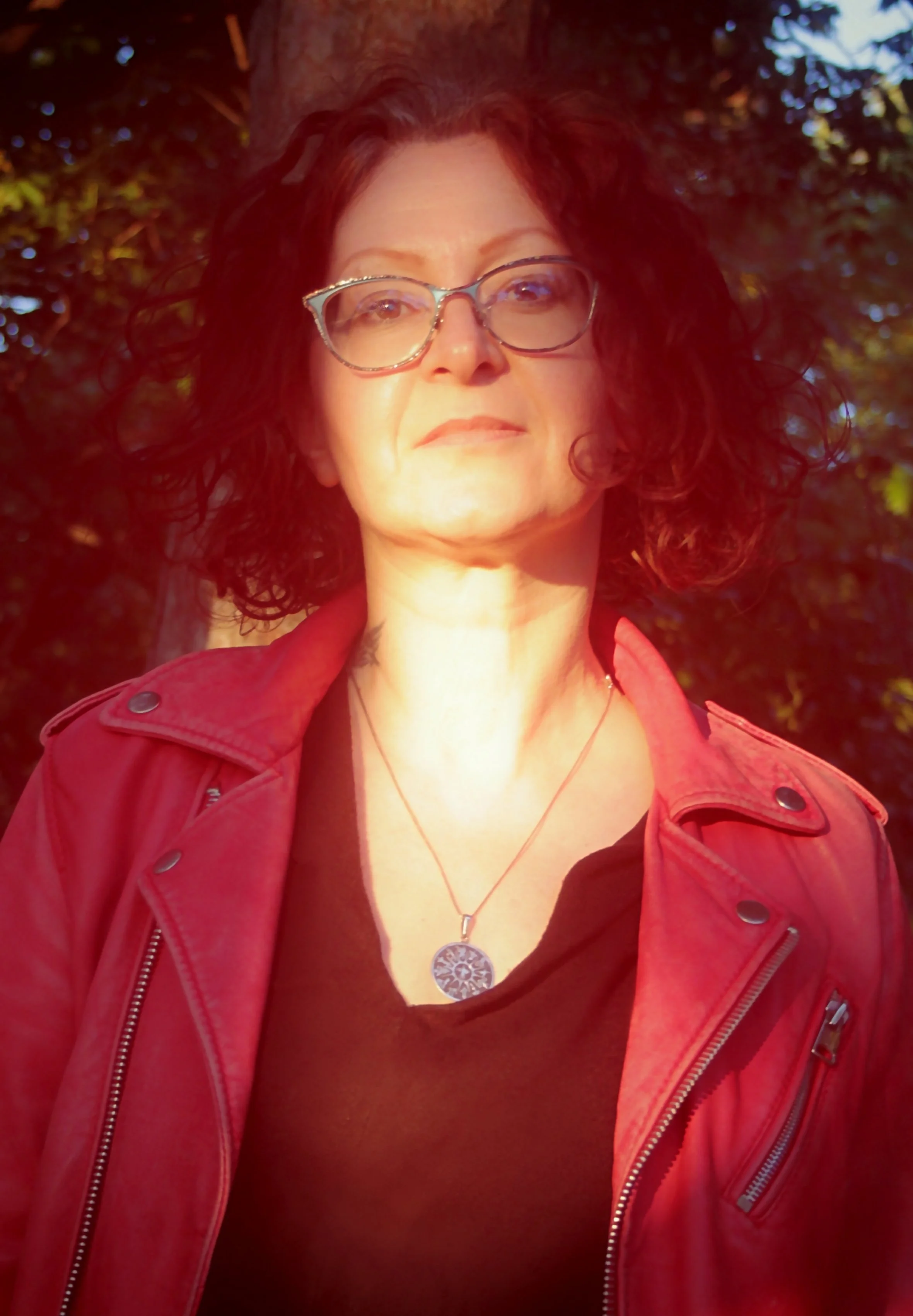 A woman with curly dark hair, glasses, and a red leather jacket standing outdoors with sunlight on her face and a background of trees.