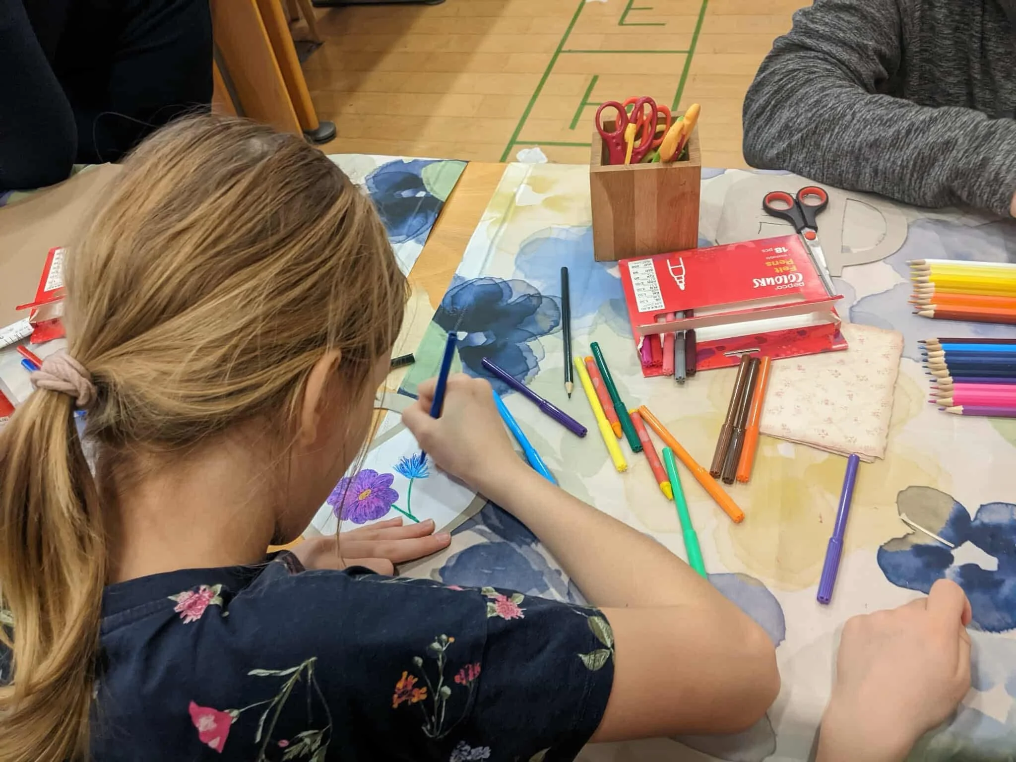 A girl with blonde hair tied in a ponytail, coloring a flower on paper with various colored markers on a table.