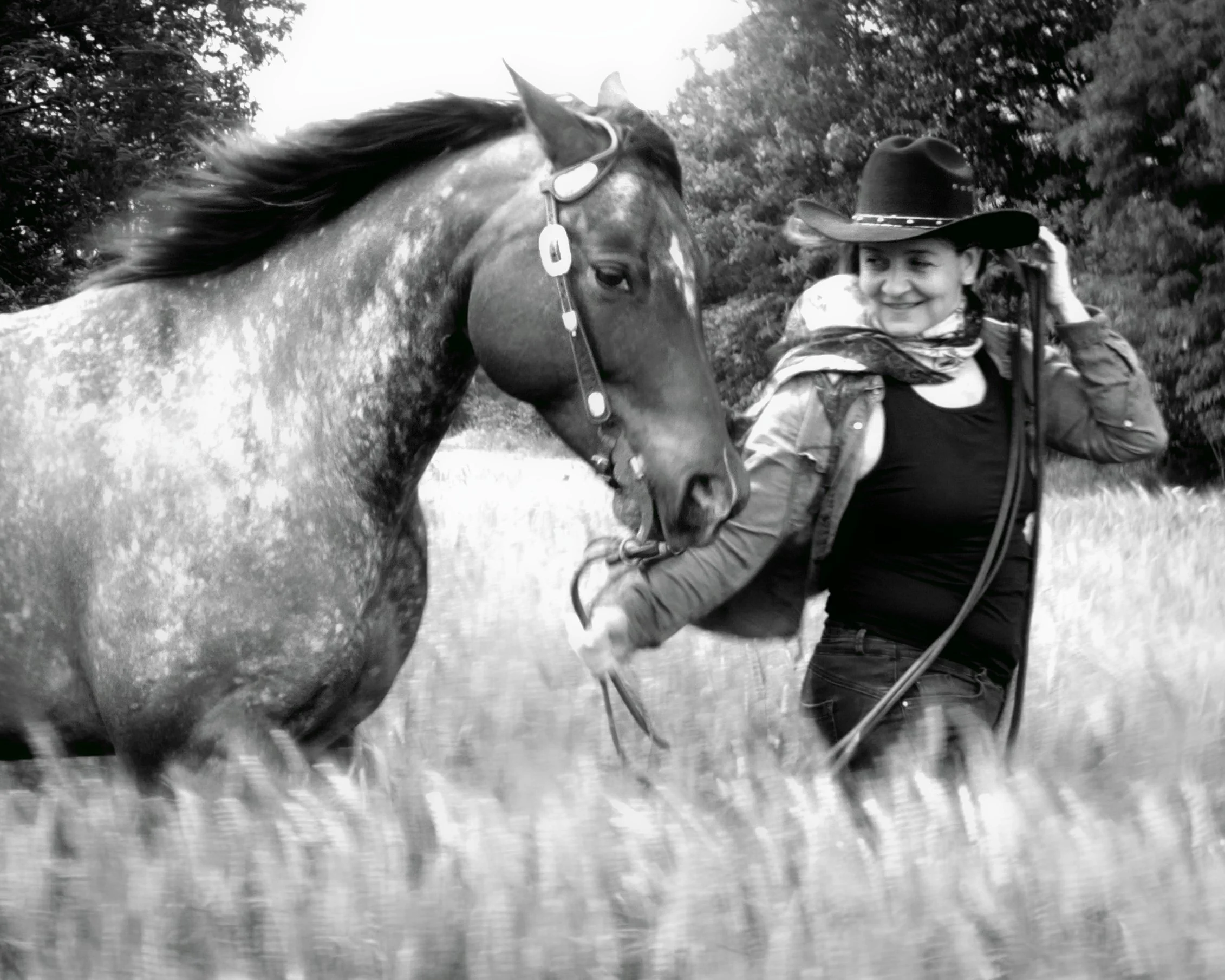 A woman with a cowboy hat smiling and looking at a horse in a field with trees in the background.