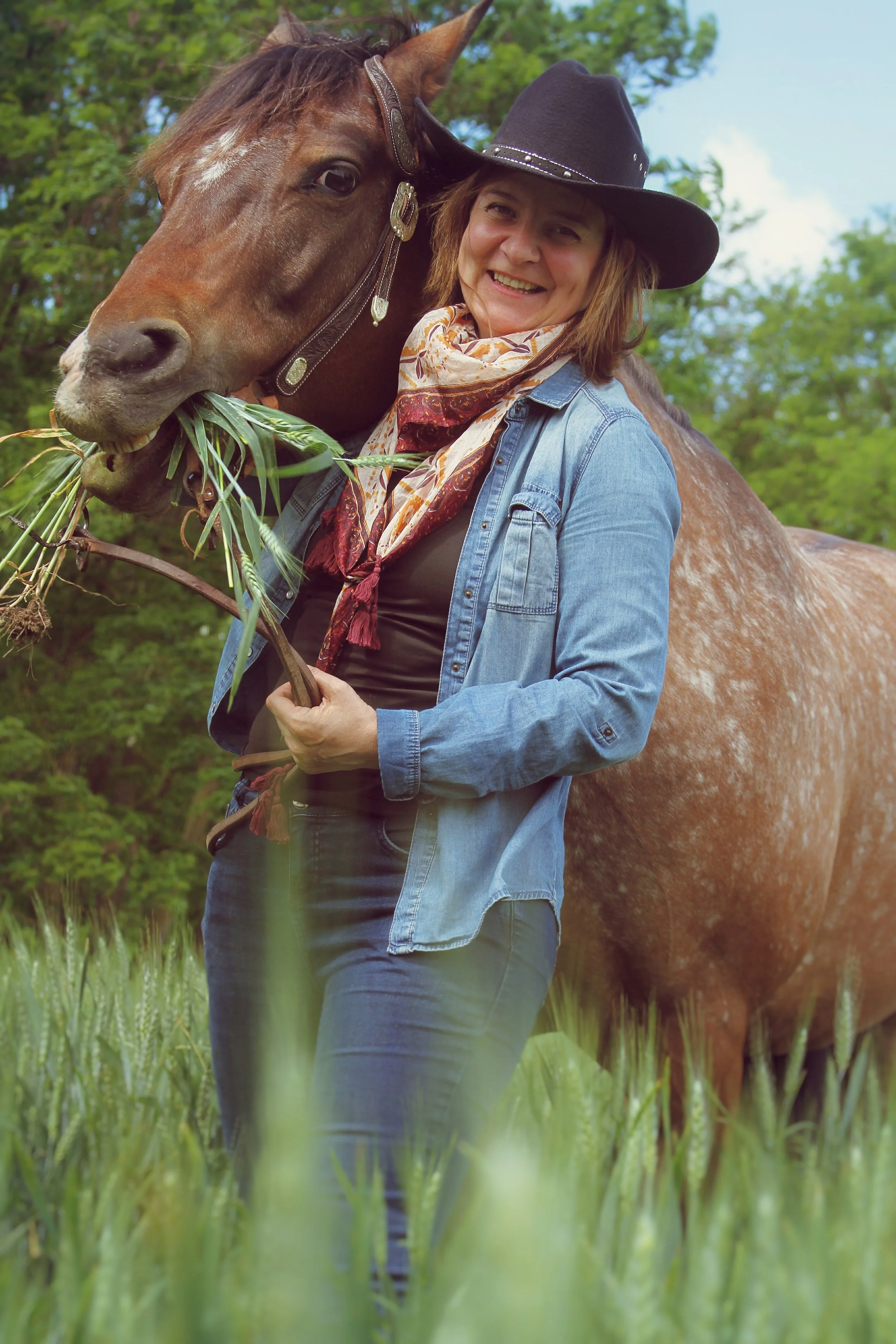 A woman in a cowboy hat and denim jacket smiling next to a horse in a field of green grass, holding a branch of grass in her hand.