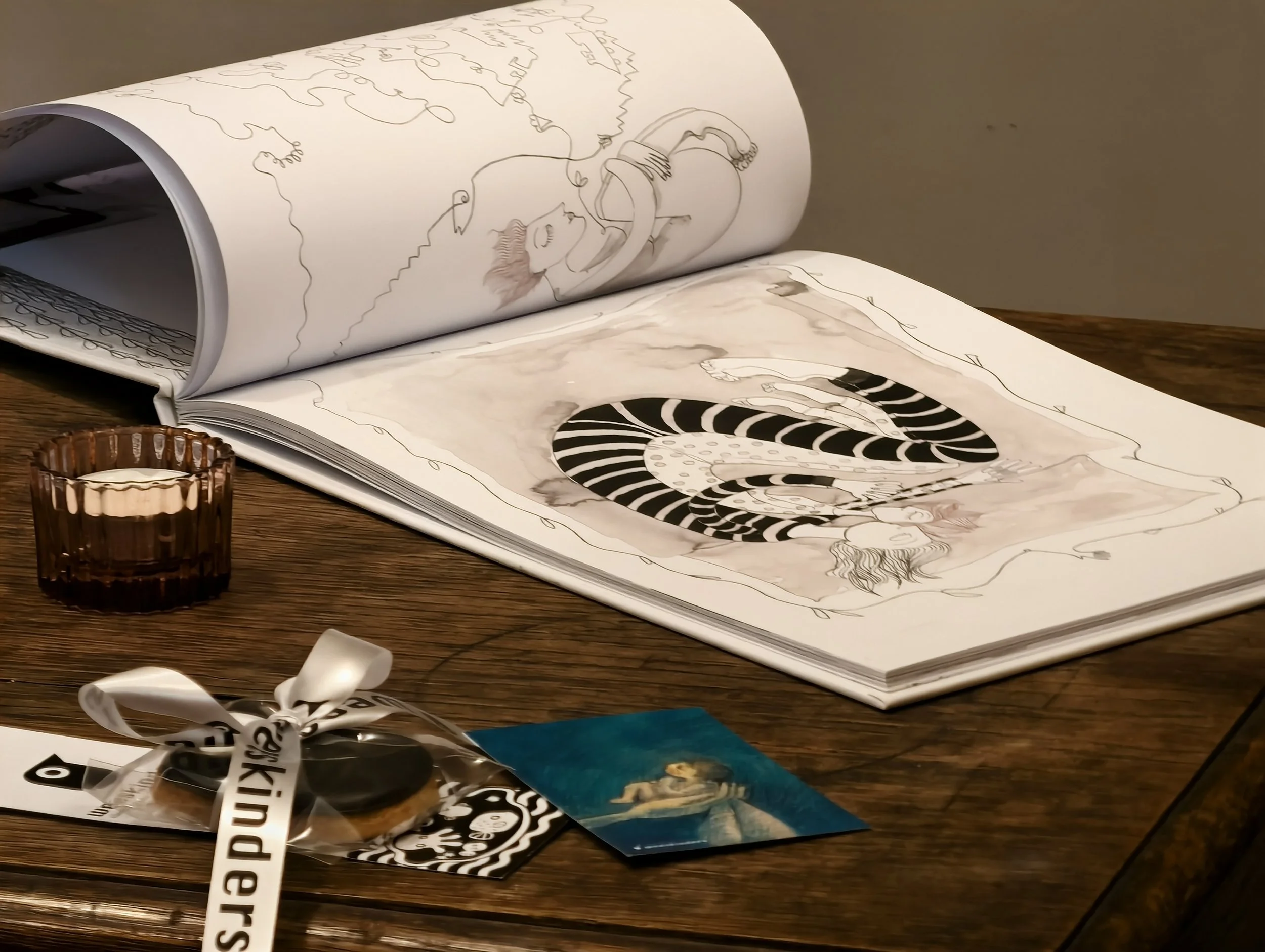 Open illustrated book with drawings of a woman and a snake on a wooden table, next to a small glass container, a wrapped cookie with a ribbon, a small picture of a woman, and a business card.