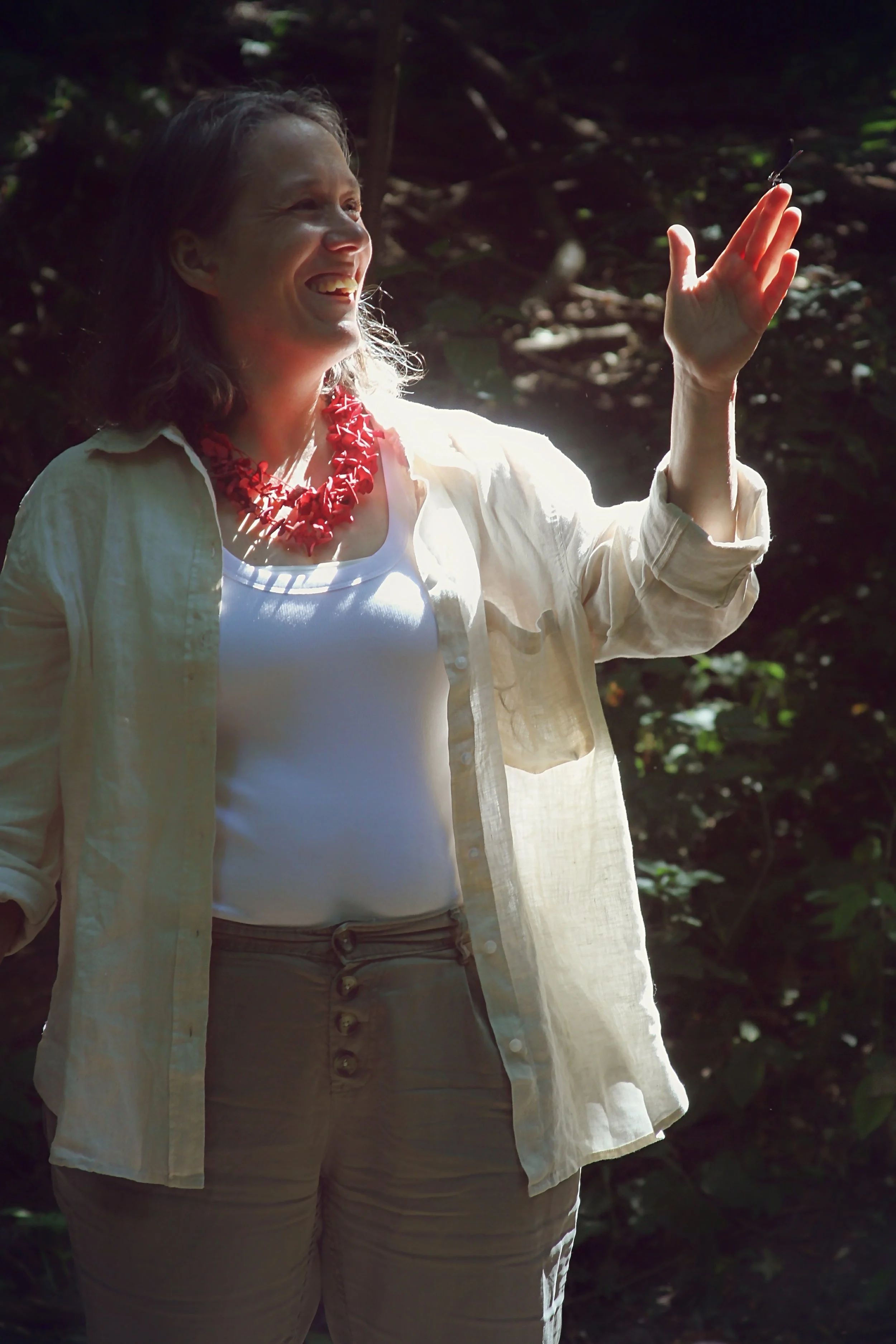 A woman with shoulder-length hair, wearing a white top, beige high-waisted pants, a cream-colored open shirt, and a red necklace, is smiling while reaching out to a butterfly in a wooded outdoor setting.