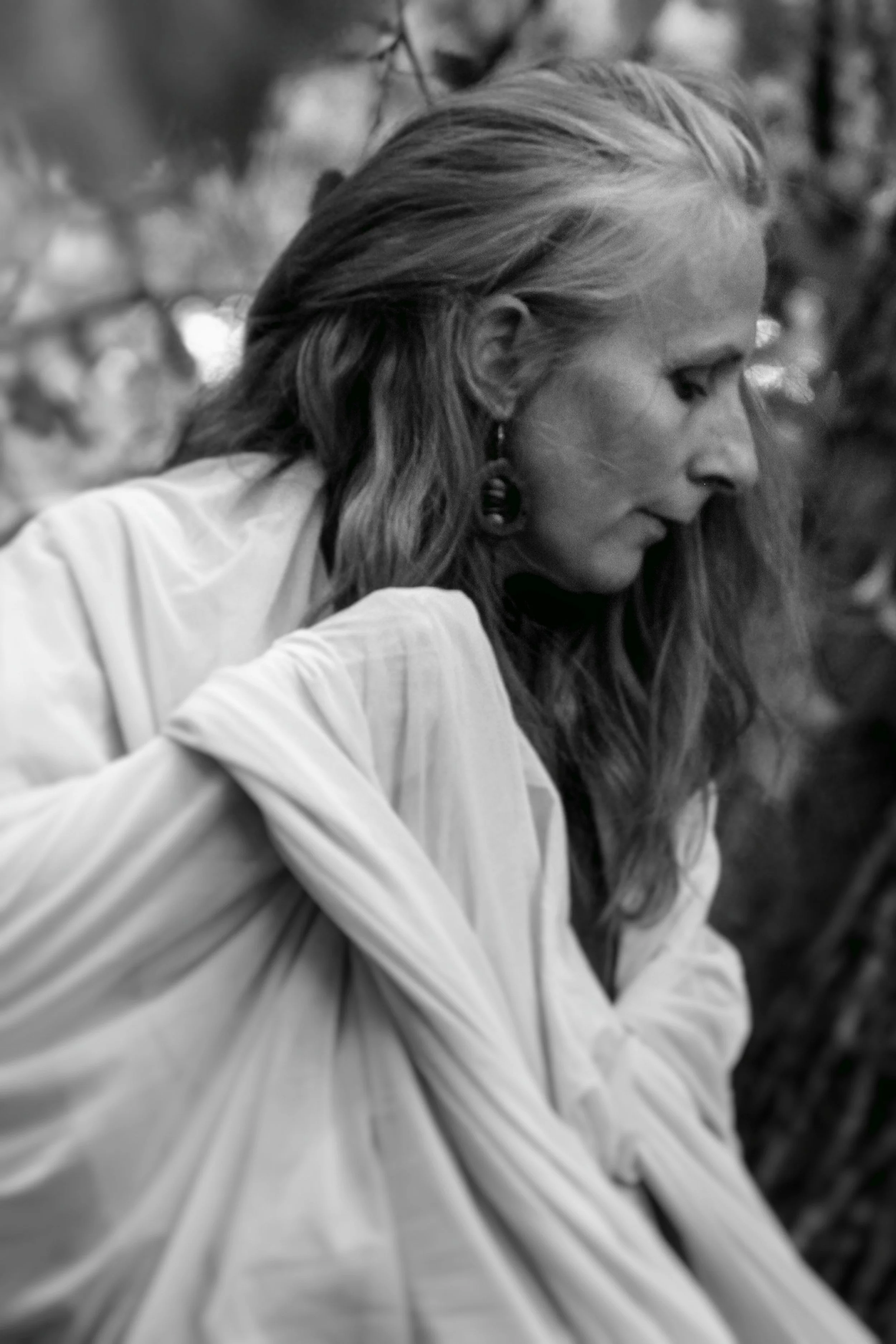 A black and white photo of a woman with long hair wearing earrings and a light-colored top, looking down with her arms crossed.