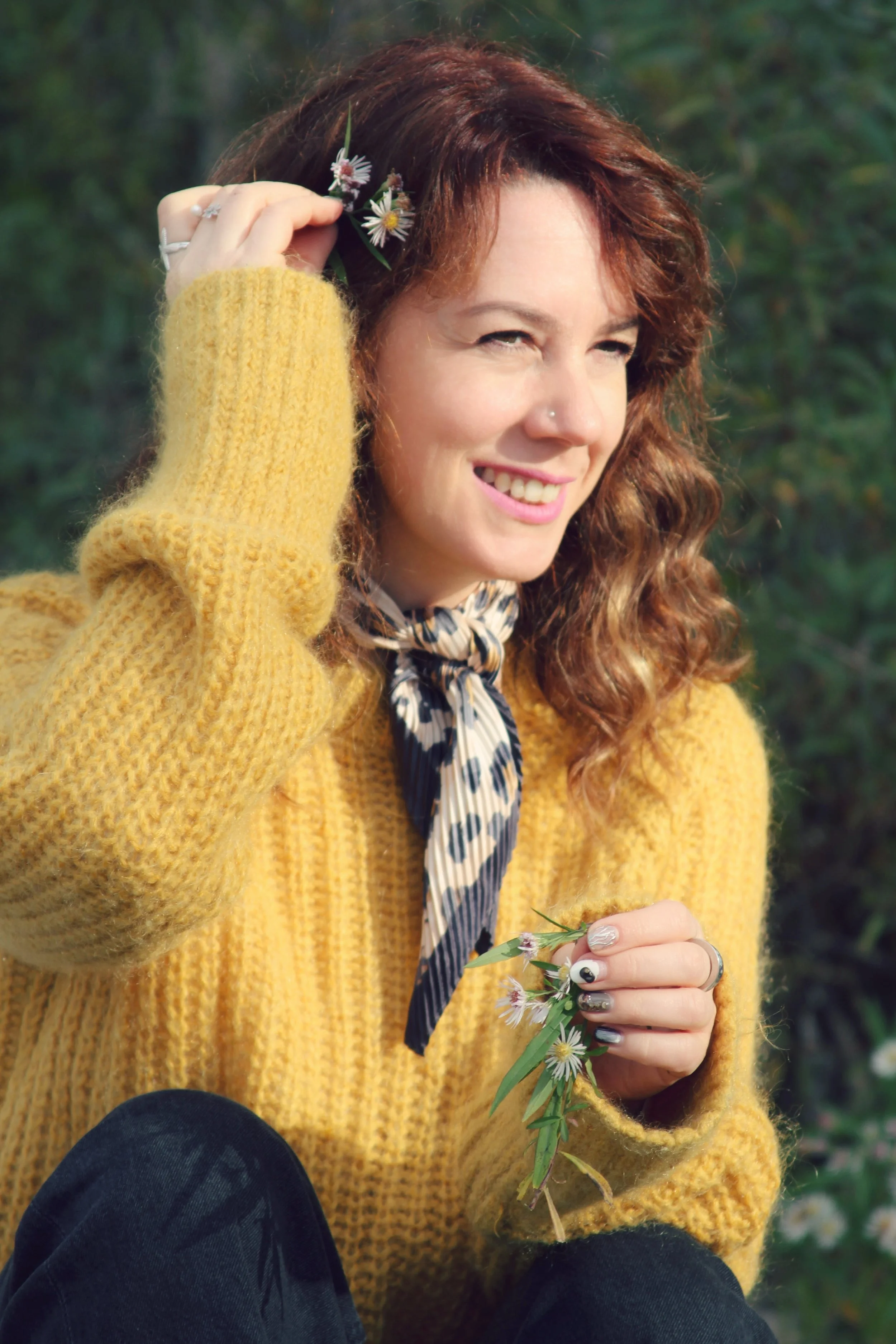 A woman sitting outdoors, smiling, wearing a yellow knitted sweater, a leopard print scarf, and holding a small bouquet of wildflowers. She has curly red hair and is holding some flowers near her ear.