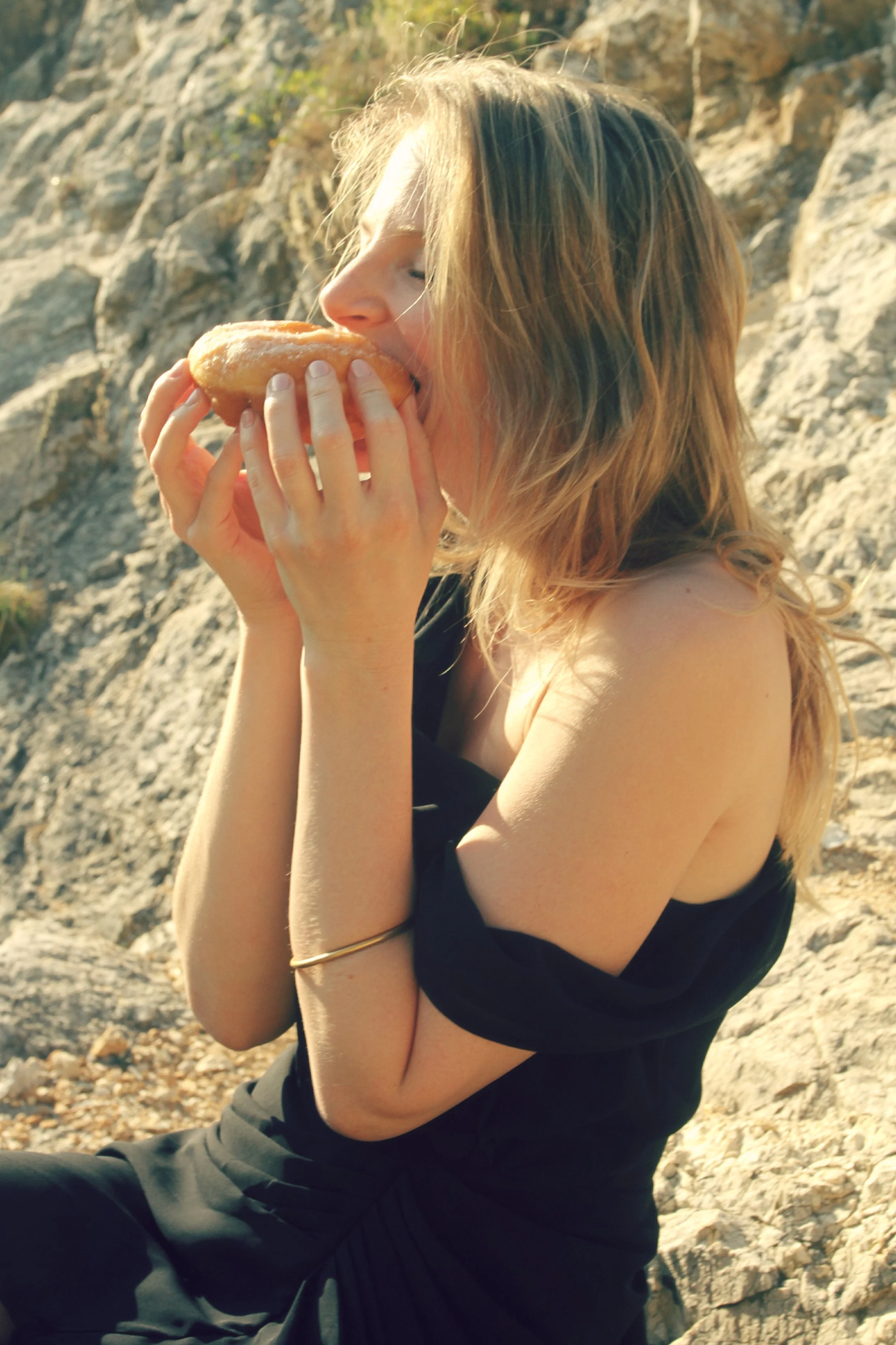 A woman with blonde hair sitting outdoors near rocky terrain, about to bite into a glazed donut.