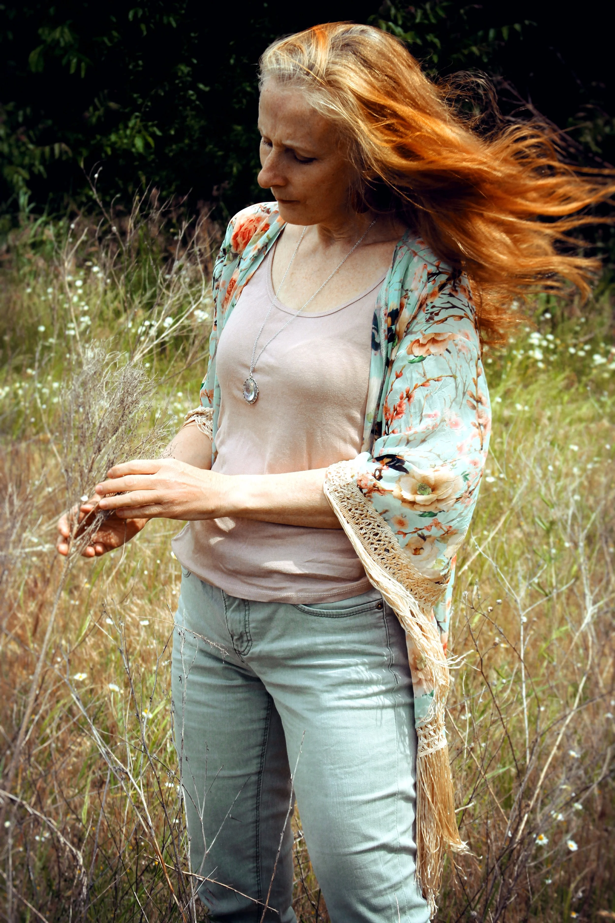 A woman with long red hair standing in a field of tall grass and wildflowers, looking down and holding a small branch.