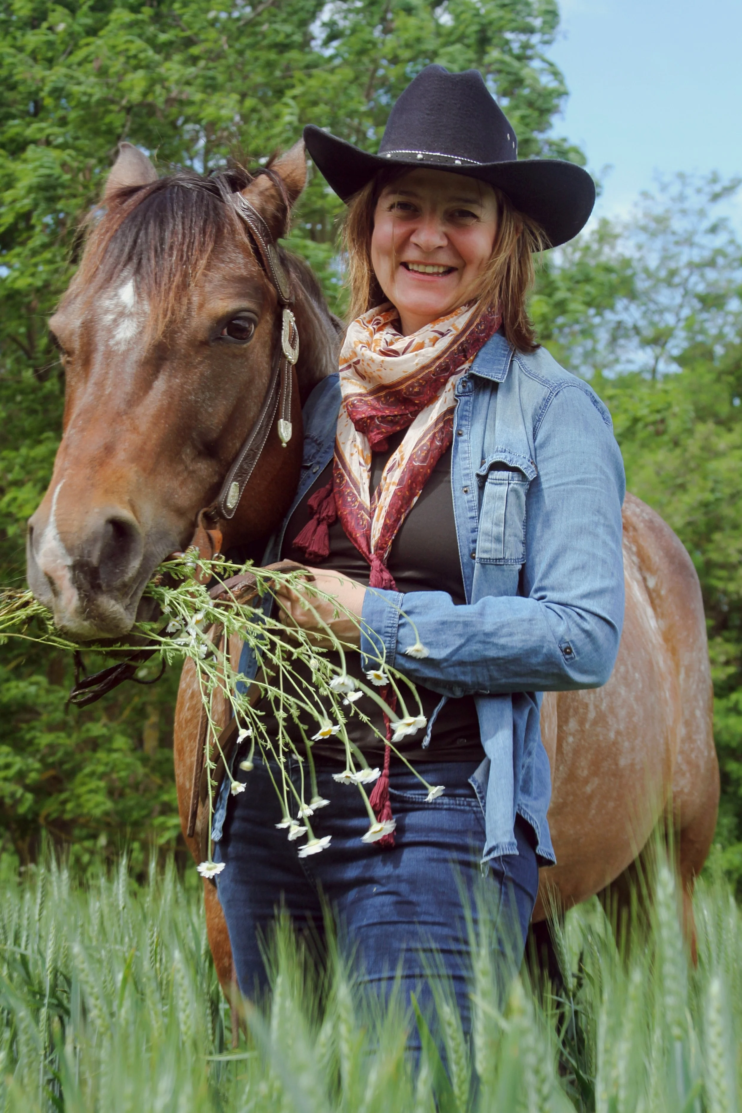 A woman with a cowboy hat, denim jacket, and scarf, smiling while standing beside a brown horse with a white star on its forehead, holding a bunch of wildflowers in her hand, in a grassy outdoor setting with trees in the background.
