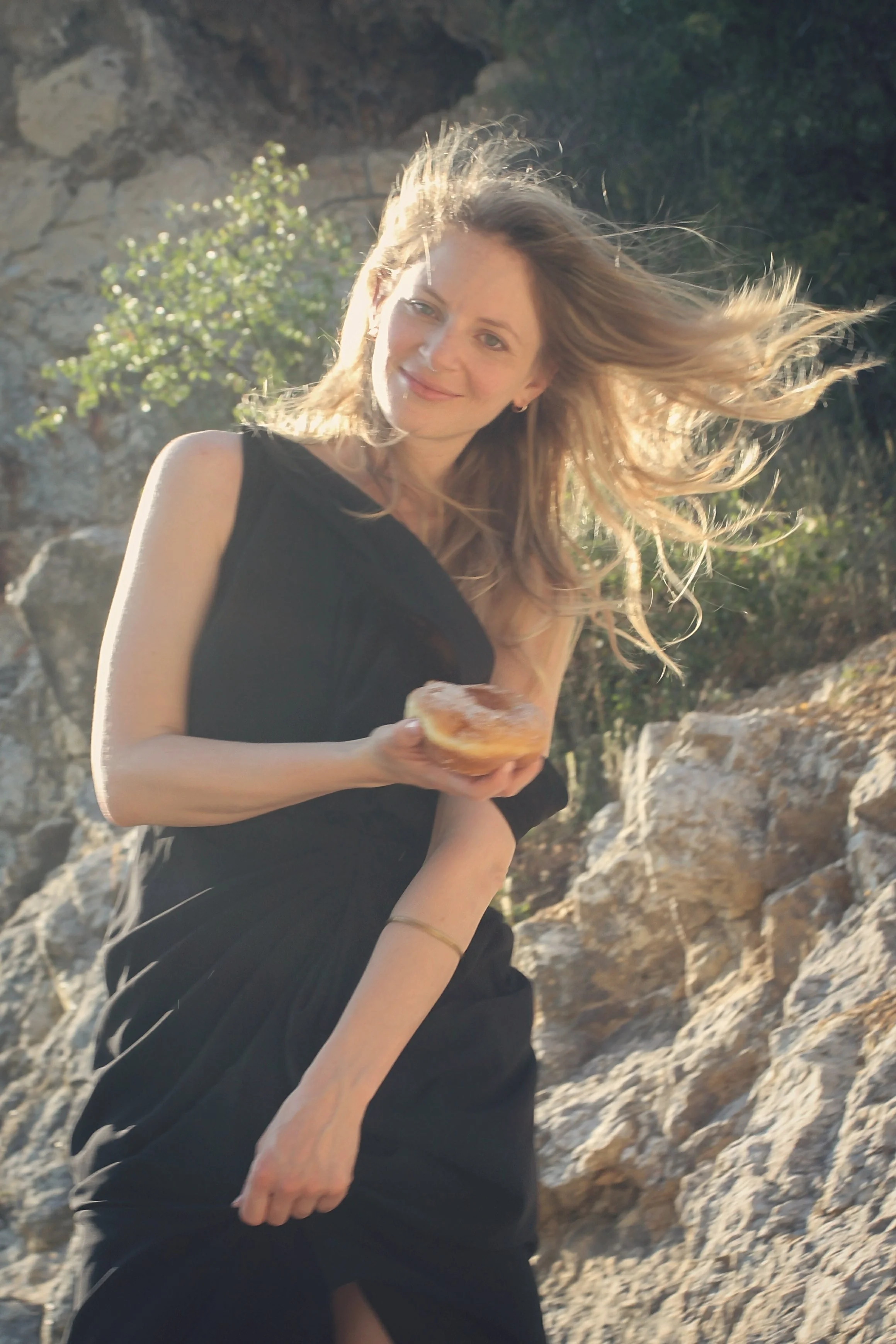 A woman with long blonde hair wearing a black dress, holding a pastry, standing outdoors among rocks and greenery, smiling at the camera.