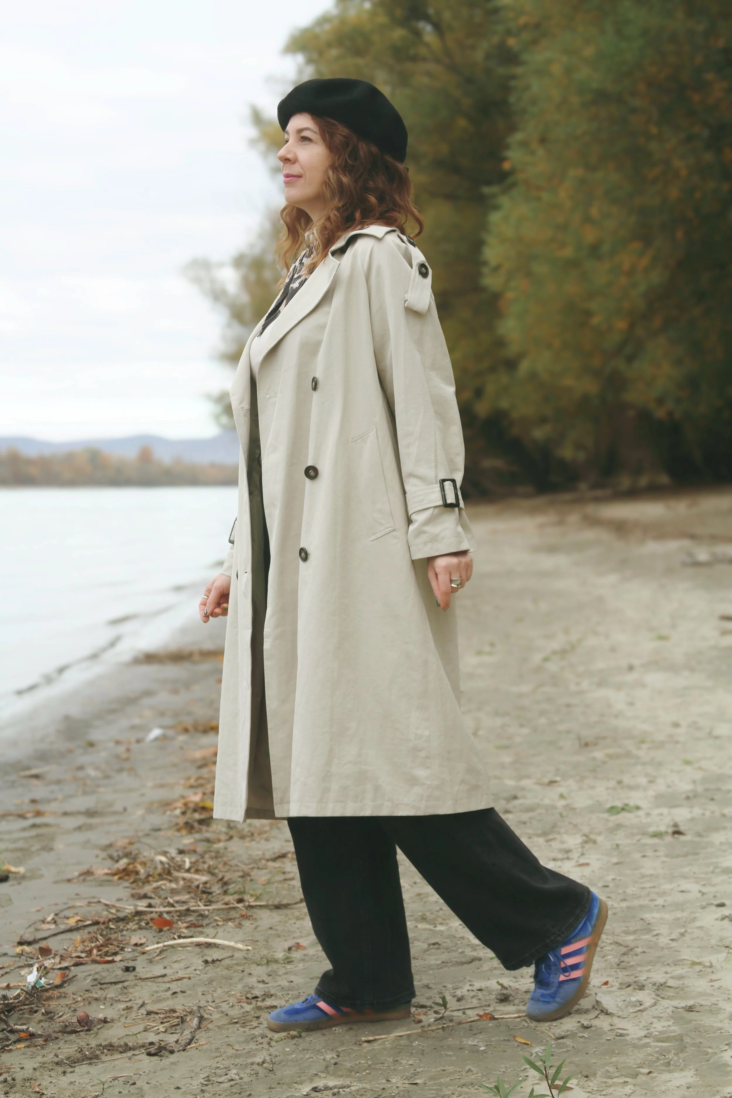 Woman standing on a beach near the water, wearing a beige trench coat, black wide-leg pants, blue sneakers with pink stripes, a black beret, and looking into the distance with trees in the background.