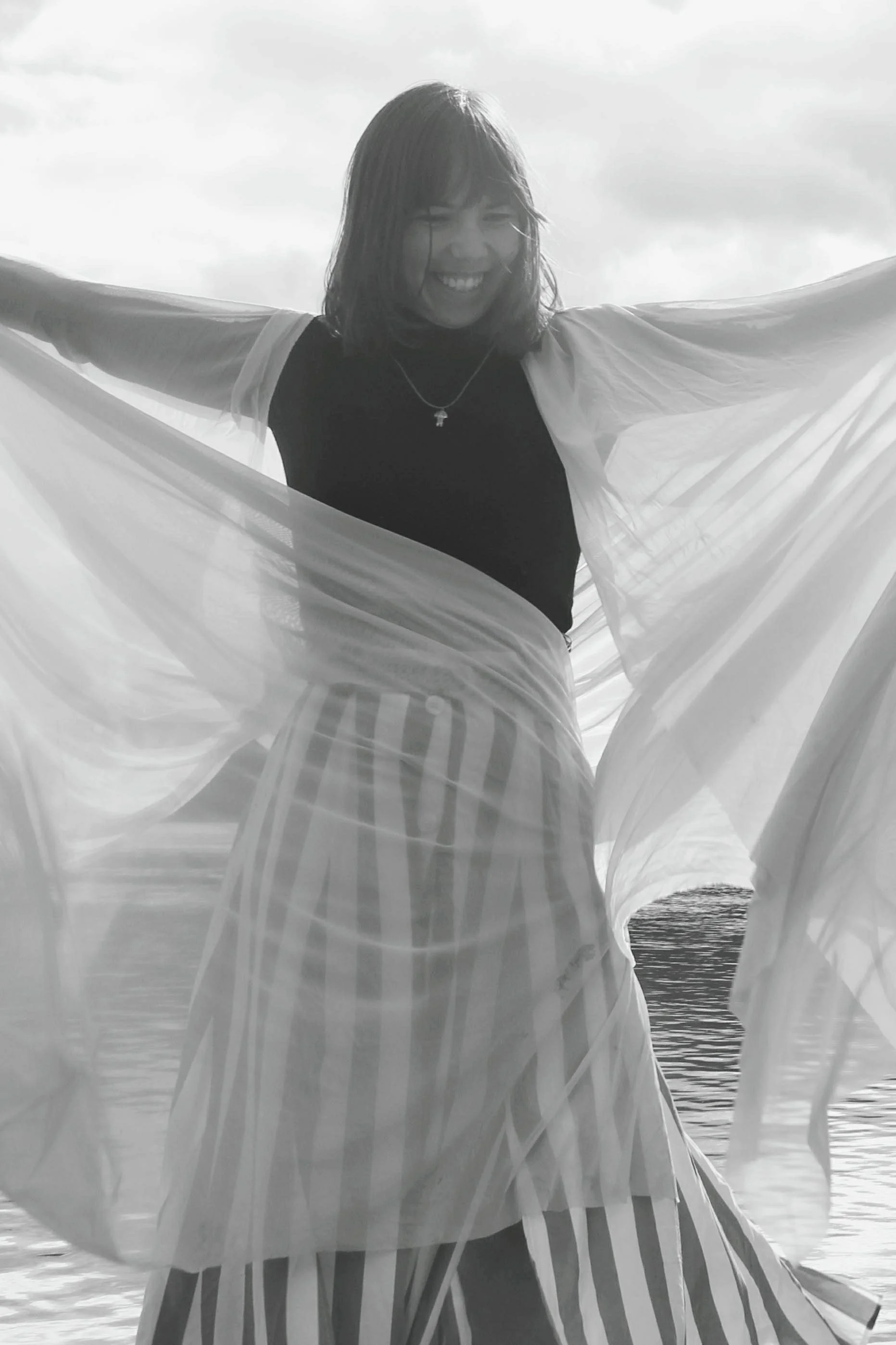 A woman smiling with arms outstretched, wearing a striped skirt and a black top, during daytime near the water with cloudy sky.