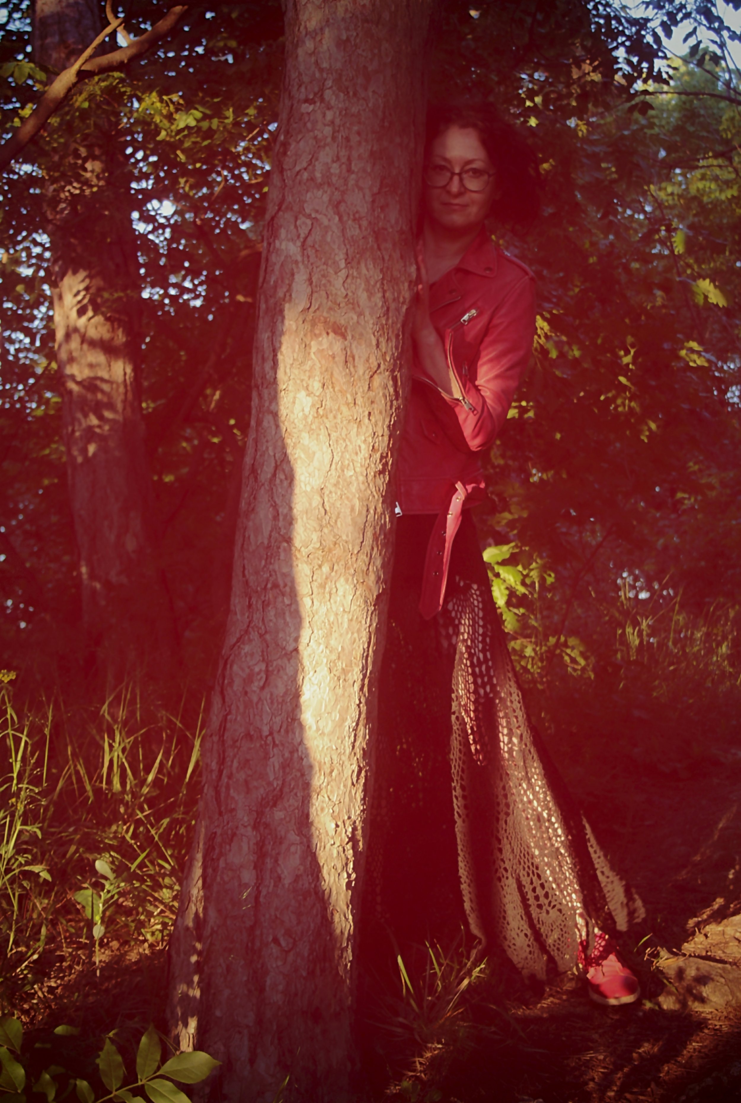 A woman with glasses leaning against a tree in a wooded area, wearing a red jacket and a long, patterned skirt.