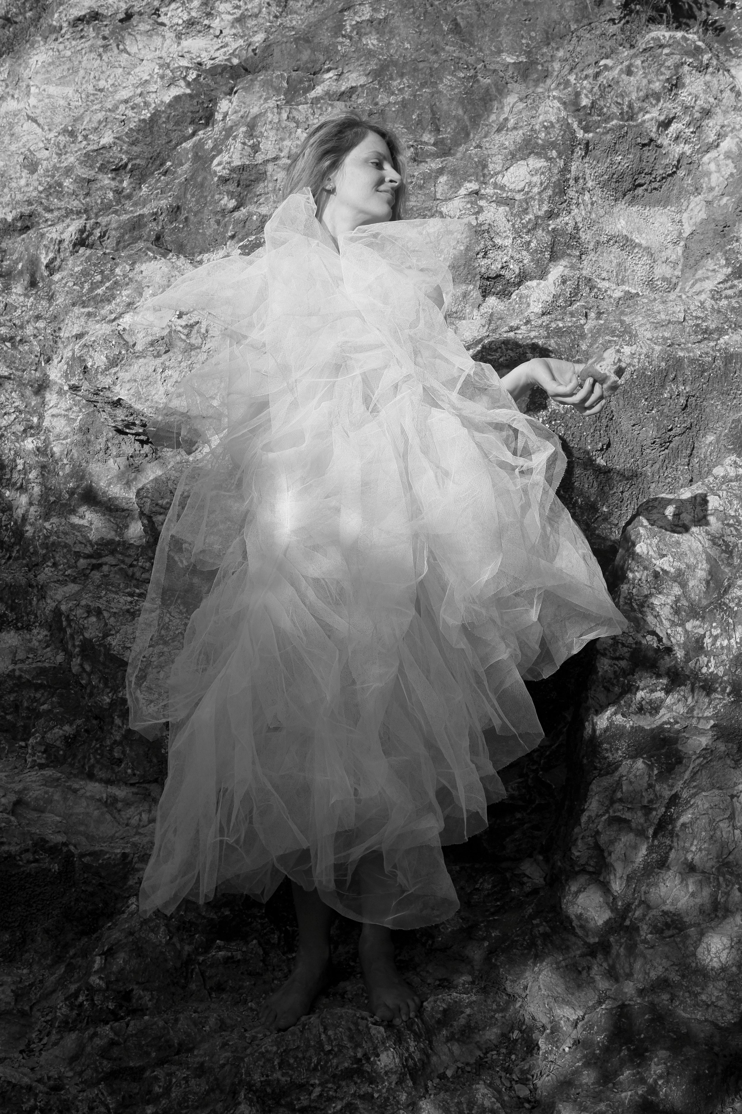 A woman in a flowing, sheer dress resting against a rocky wall outdoors.