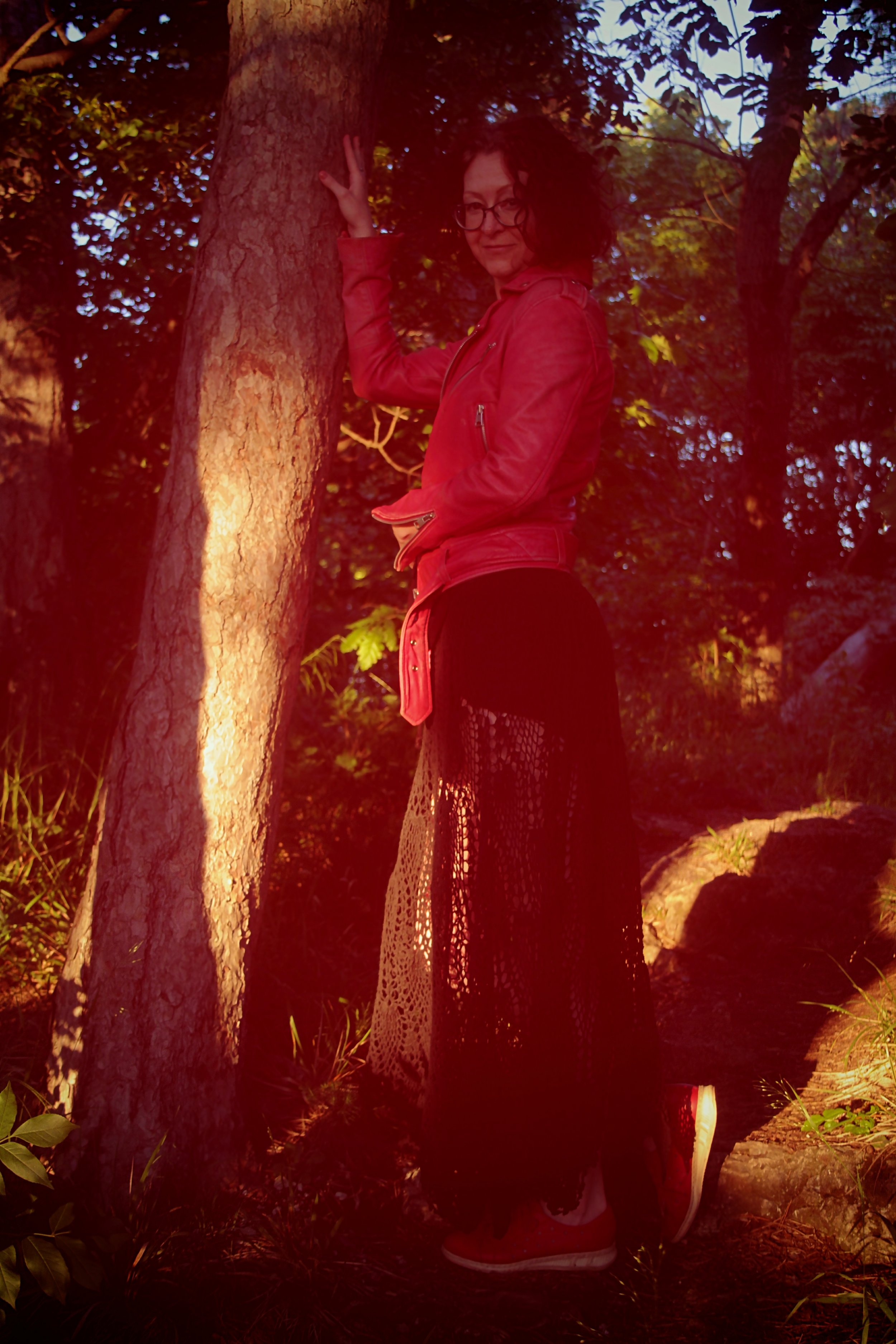 A woman with curly hair and glasses wearing a red jacket and black lace skirt standing next to a tree in a wooded area during sunset.