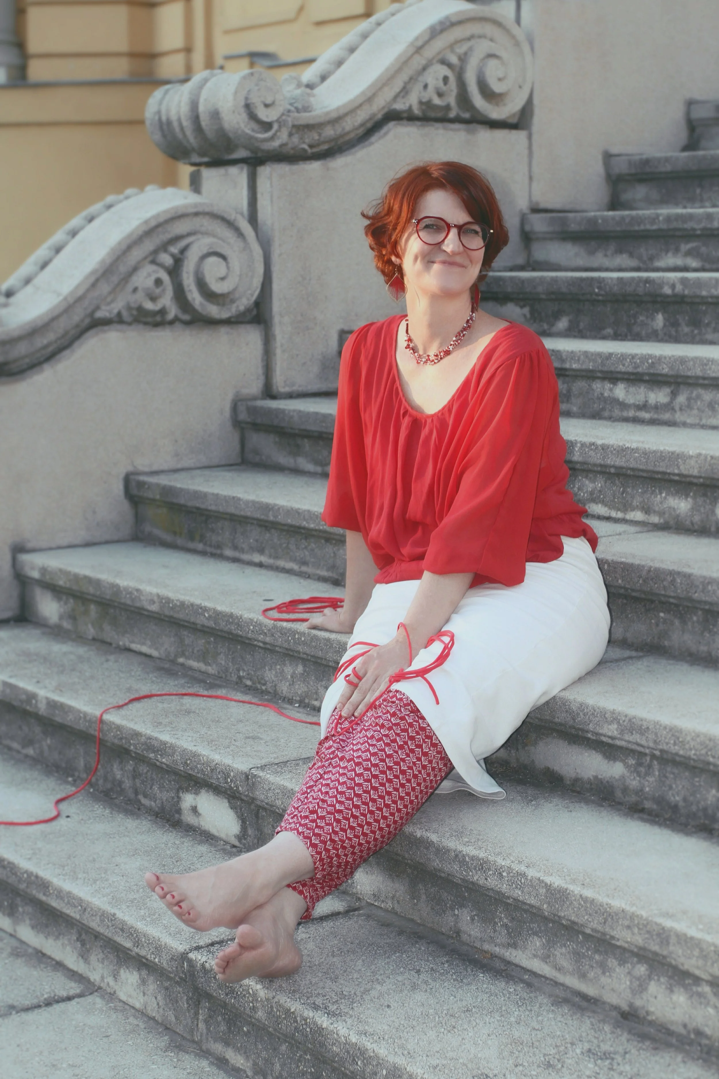 Woman sitting barefoot on stone stairs, dressed in a red blouse and patterned red and white pants, smiling, with ornate stone balustrades in the background.