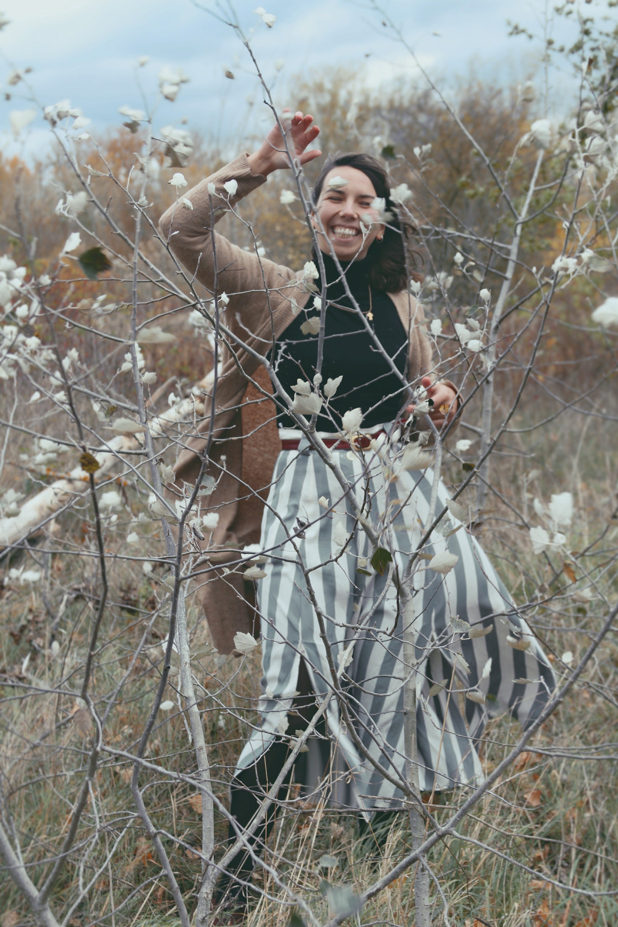 A woman smiles and laughs while standing behind a small, leafless tree with white flowers in a field during autumn, with other trees and a cloudy sky in the background.