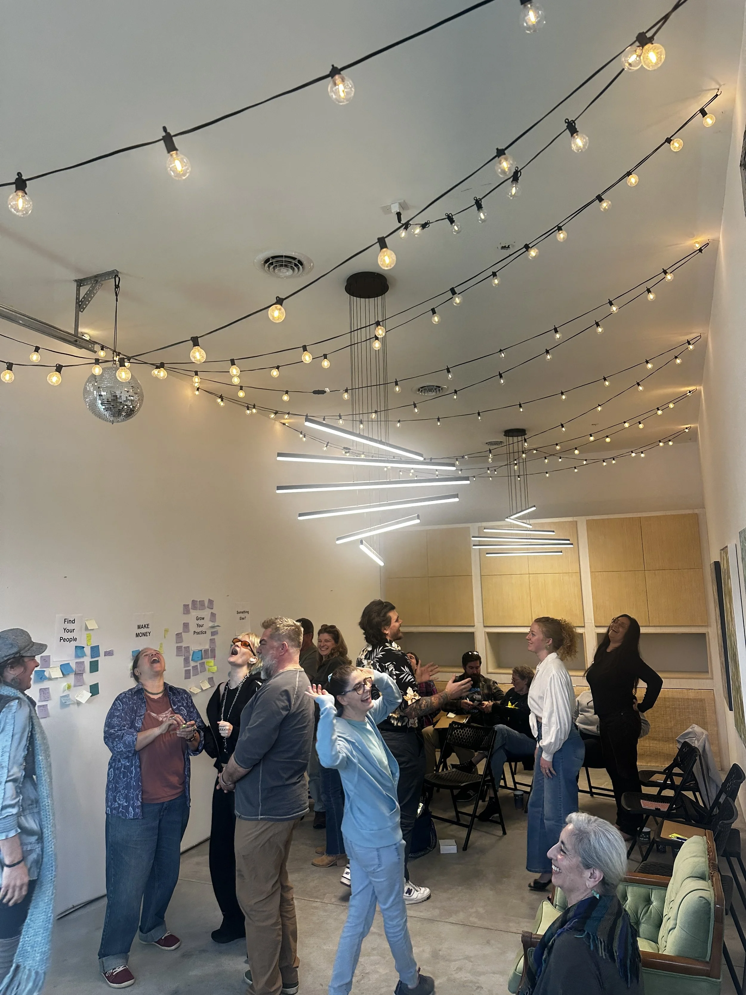 A group of people in a cozy room with colorful, abstract wall art, gathered around a wooden coffee table. Some are sitting on couches while others stand behind, smiling and posing for the photo. The table holds magazines and a drink cup. The atmosphere appears relaxed and friendly.