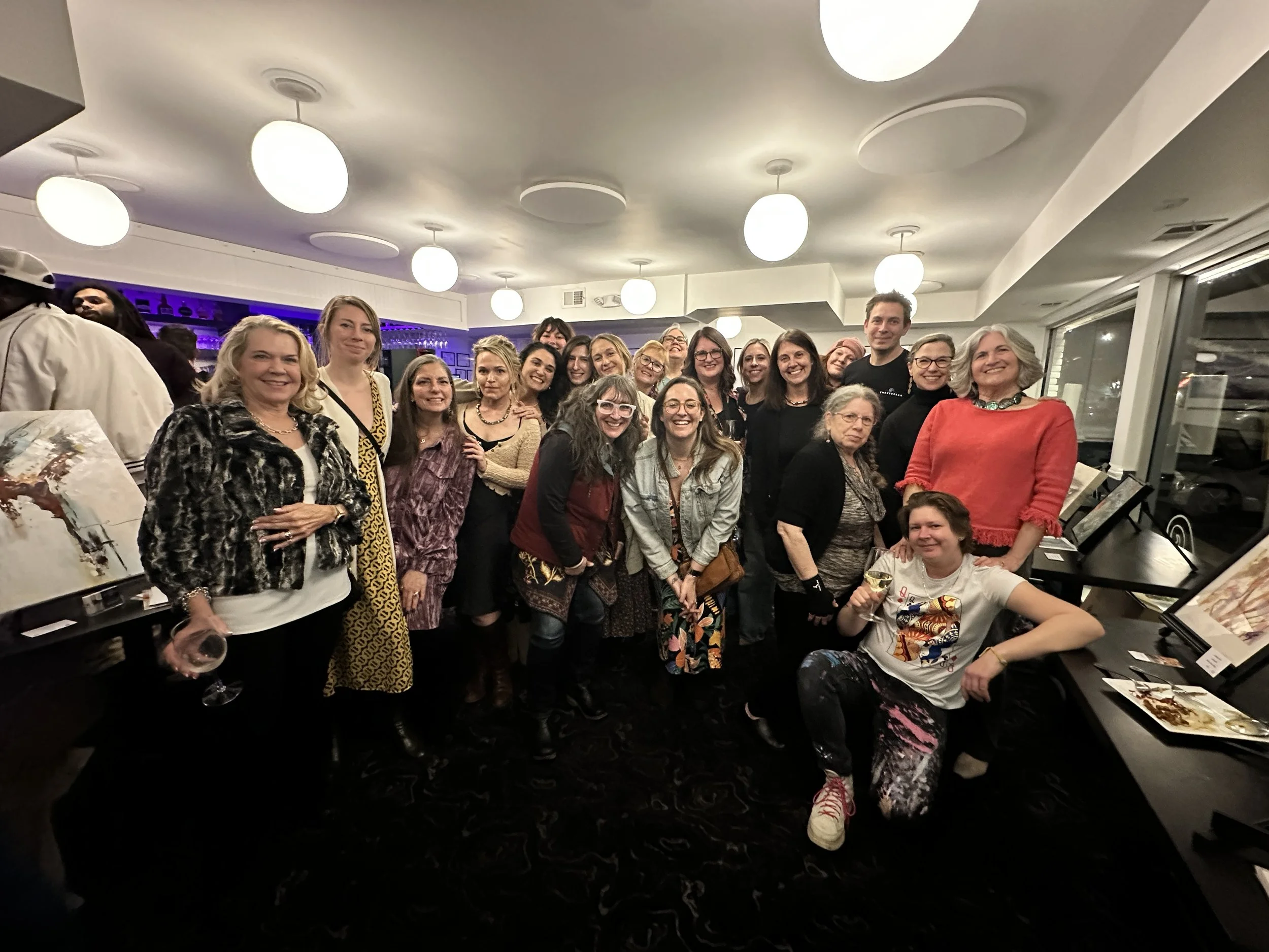 A group of people gathered in a well-lit room with circular ceiling lights, smiling for a group photo. Some artwork is displayed on tables nearby, and a person holds a wine glass. The setting appears social and festive.