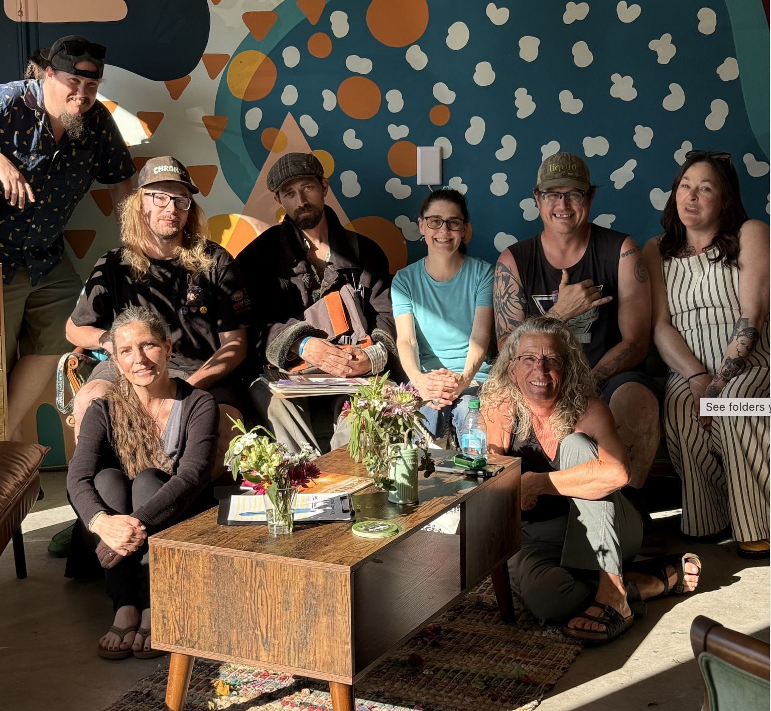A group of people in a cozy room with colorful, abstract wall art, gathered around a wooden coffee table. Some are sitting on couches while others stand behind, smiling and posing for the photo. The table holds magazines and a drink cup. The atmosphere appears relaxed and friendly.