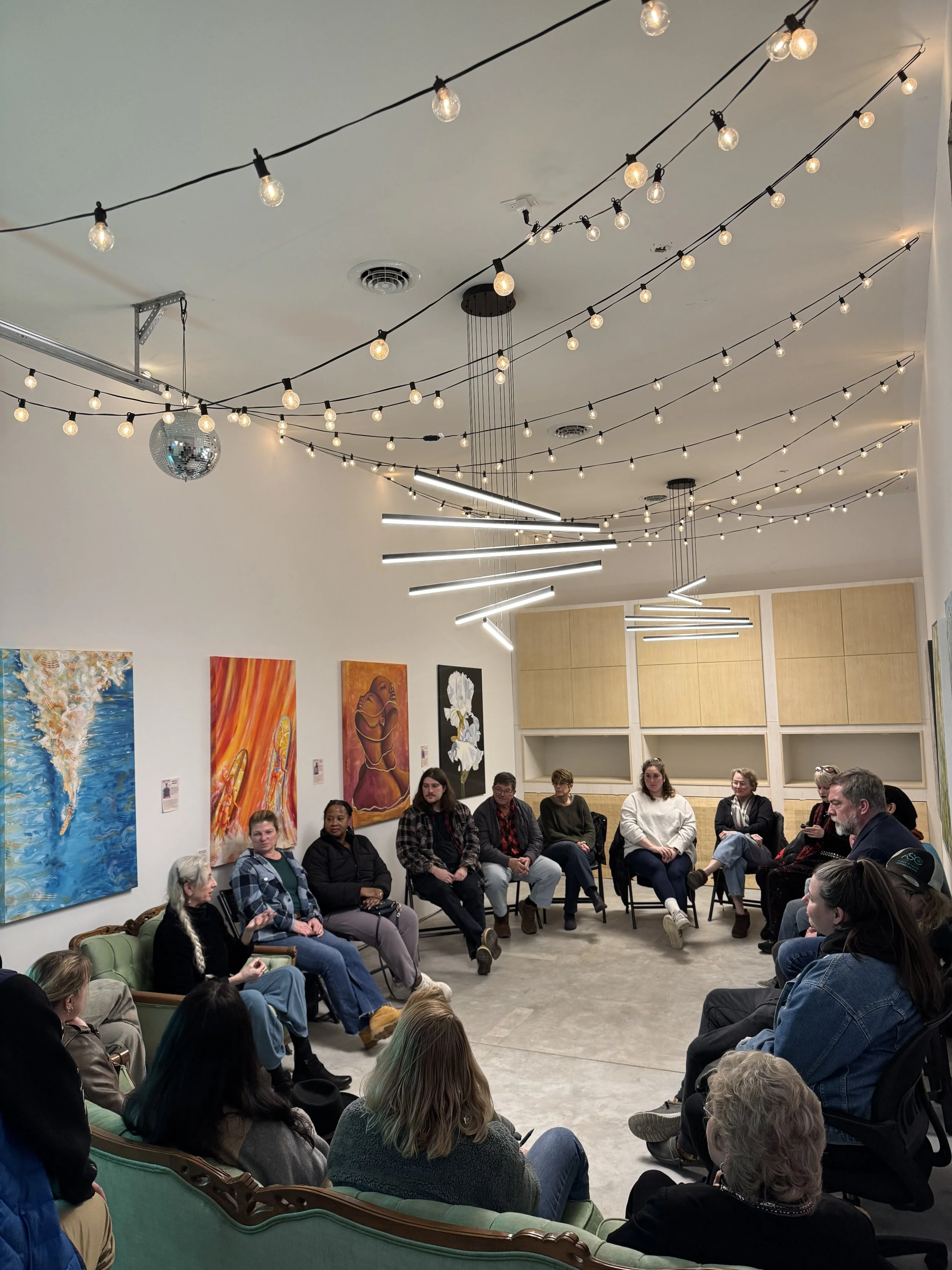 A group of people in a cozy room with colorful, abstract wall art, gathered around a wooden coffee table. Some are sitting on couches while others stand behind, smiling and posing for the photo. The table holds magazines and a drink cup. The atmosphere appears relaxed and friendly.