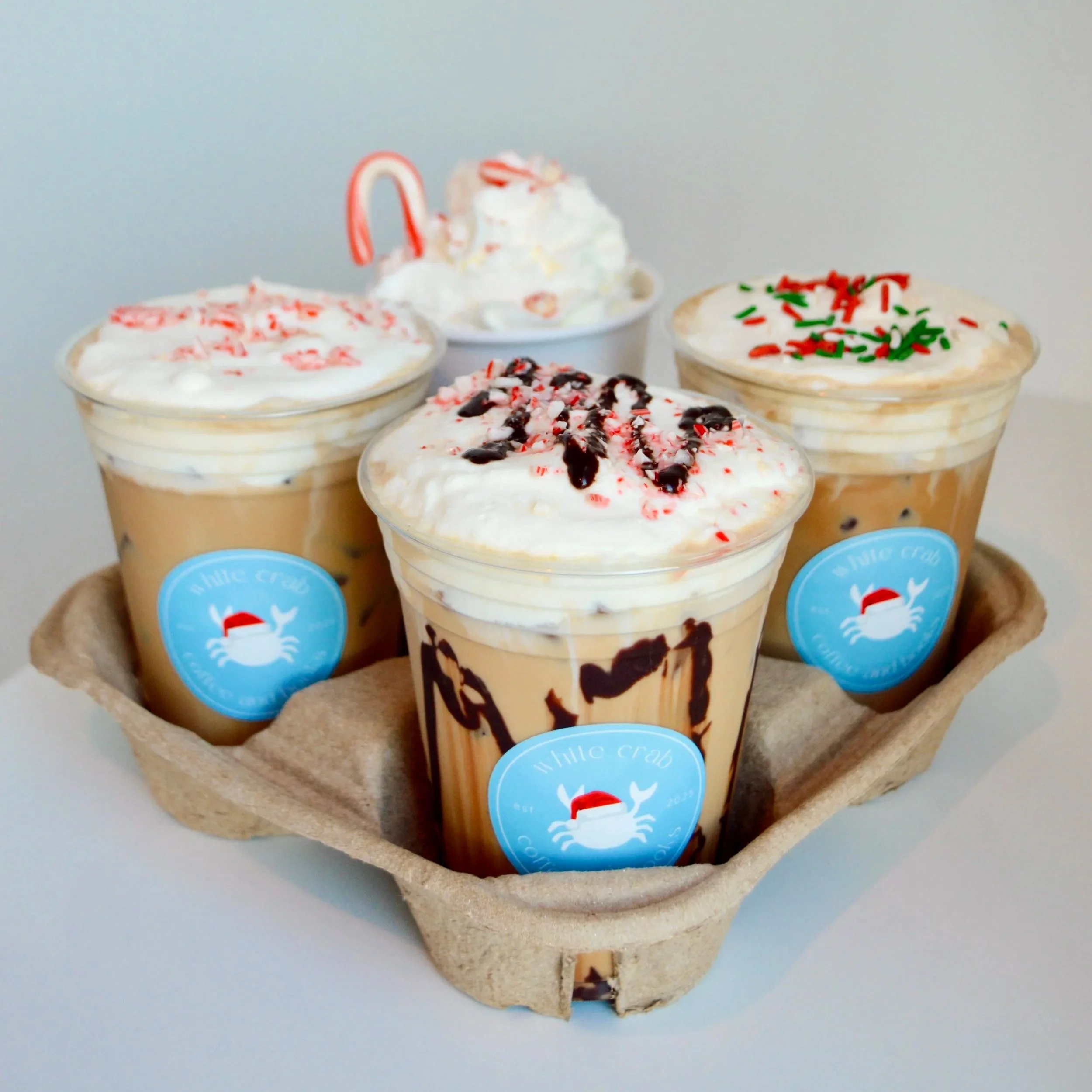 Four cups of iced coffee with whipped cream and holiday sprinkles or syrup, placed on a cardboard tray.