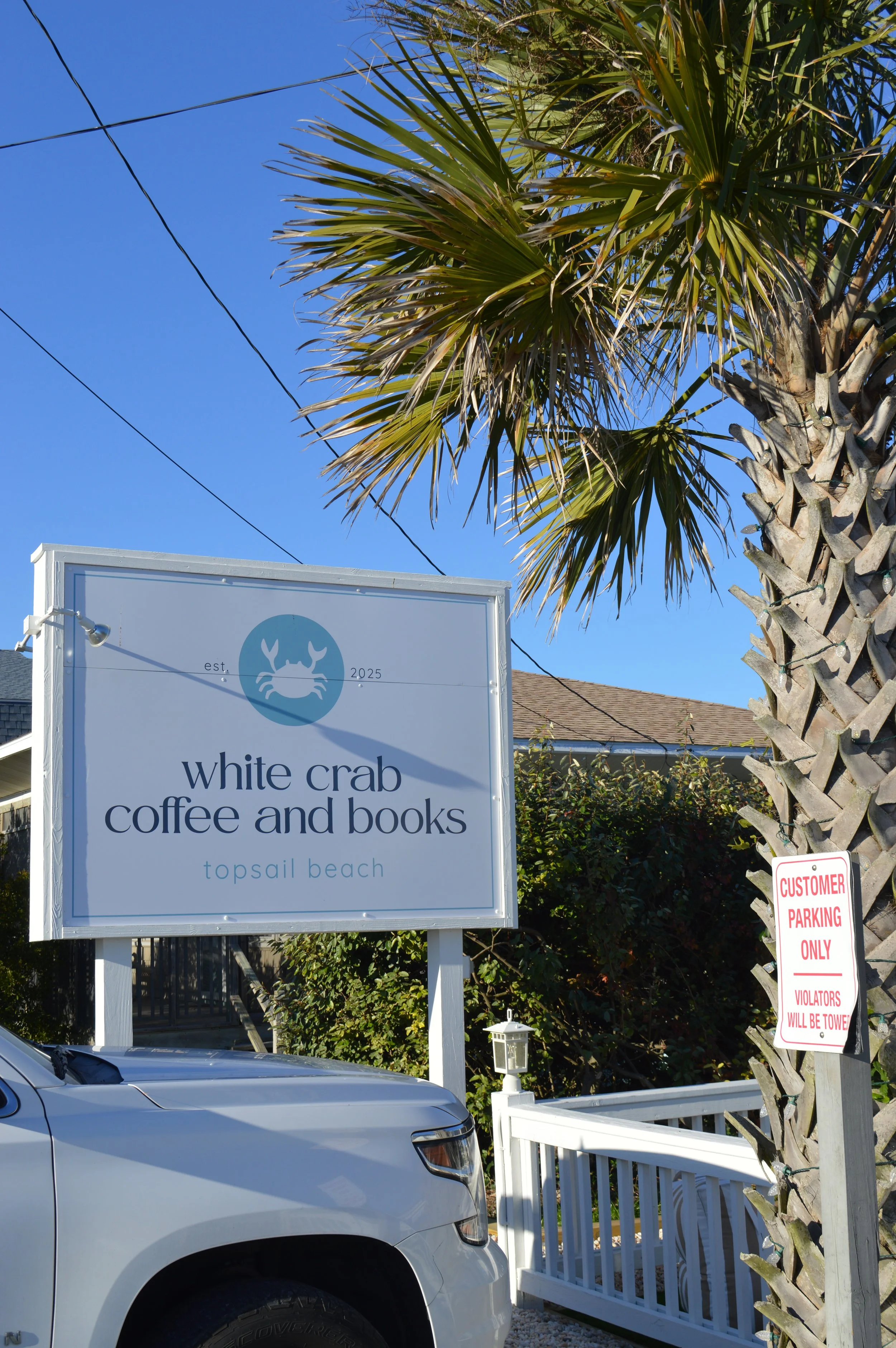 Sign for White Crab Coffee and Books at Topsail Beach, with a palm tree and a parked white vehicle nearby. A smaller sign indicates customer parking only.