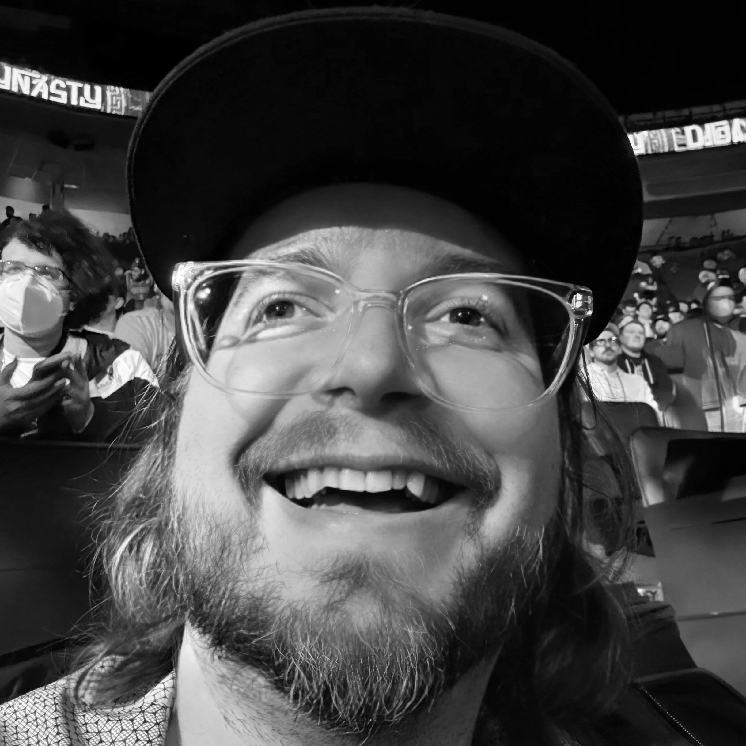 A person wearing glasses and a baseball cap, smiling. The photo appears to be upside down, and there are people in the background, some wearing masks, in what appears to be an indoor setting with a crowd.