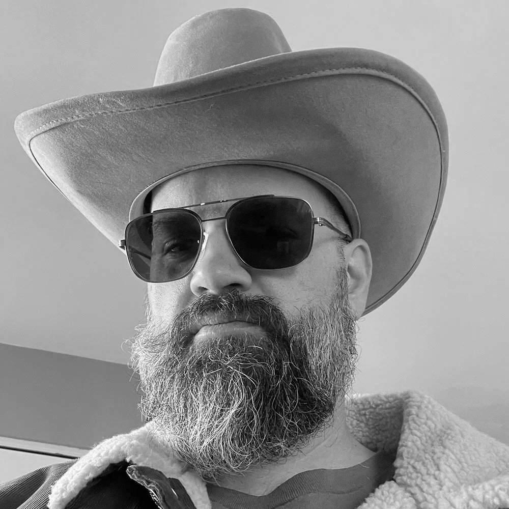 A man with a beard wearing sunglasses, a wide-brimmed hat, and a jacket with a fleece collar, looking directly at the camera.