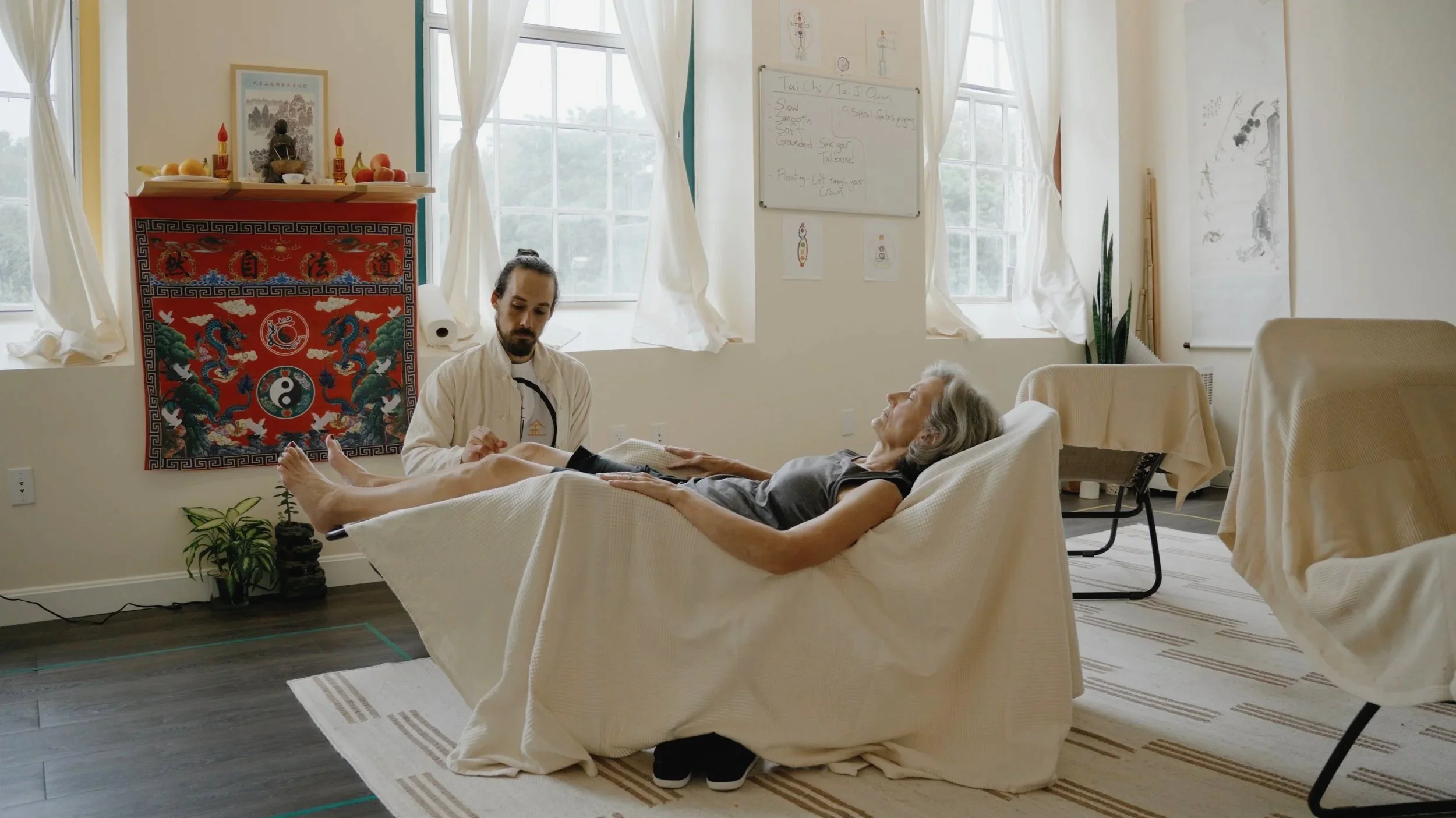 A person lying on a treatment table during an acupuncture or holistic healing session, with a practitioner attending to them in a well-lit room decorated with Asian art and plants.