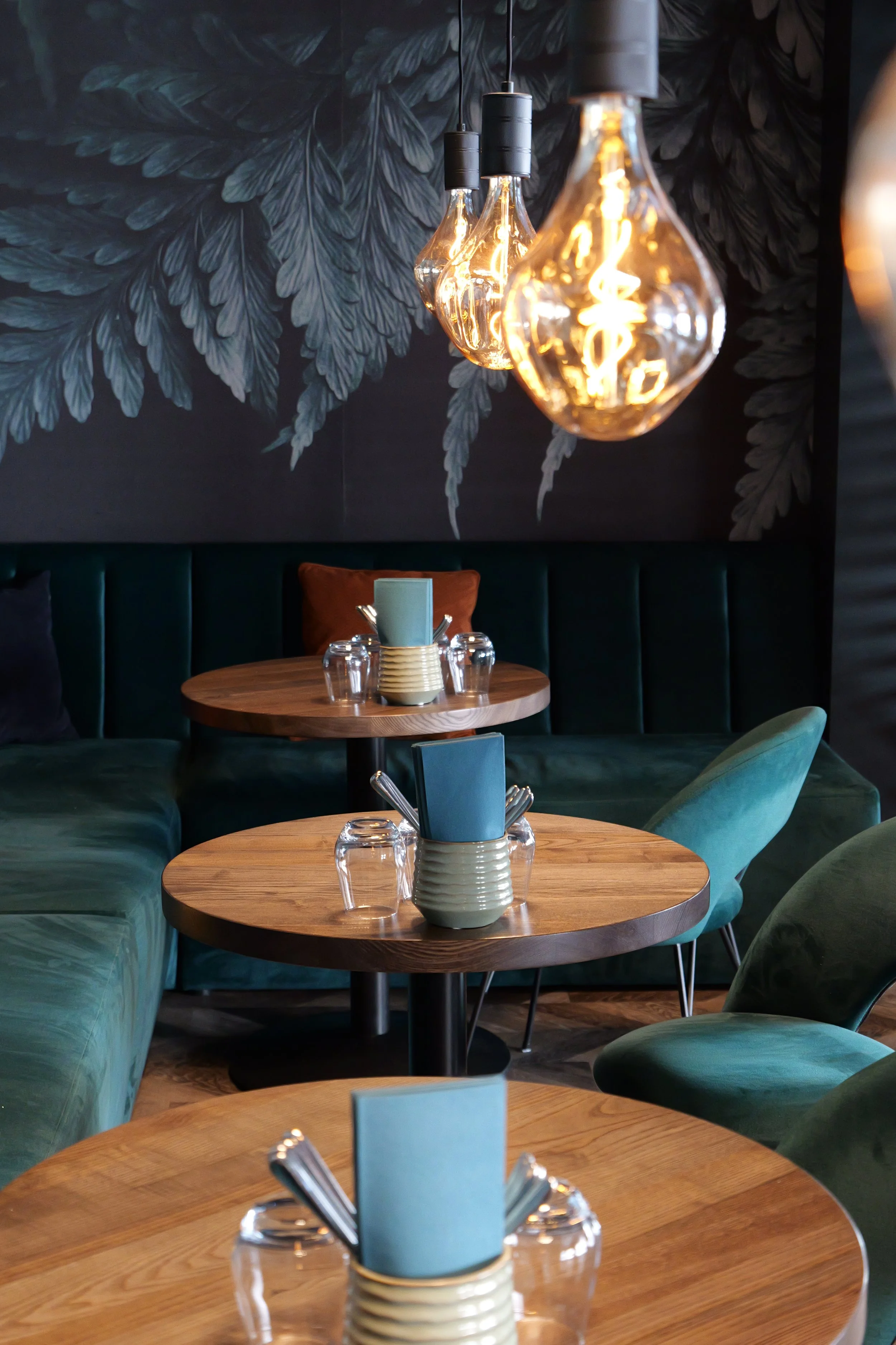 Interior of a modern cafe with wooden tables, green velvet chairs, pendant lights, and a dark wall with a round mirror and menu board