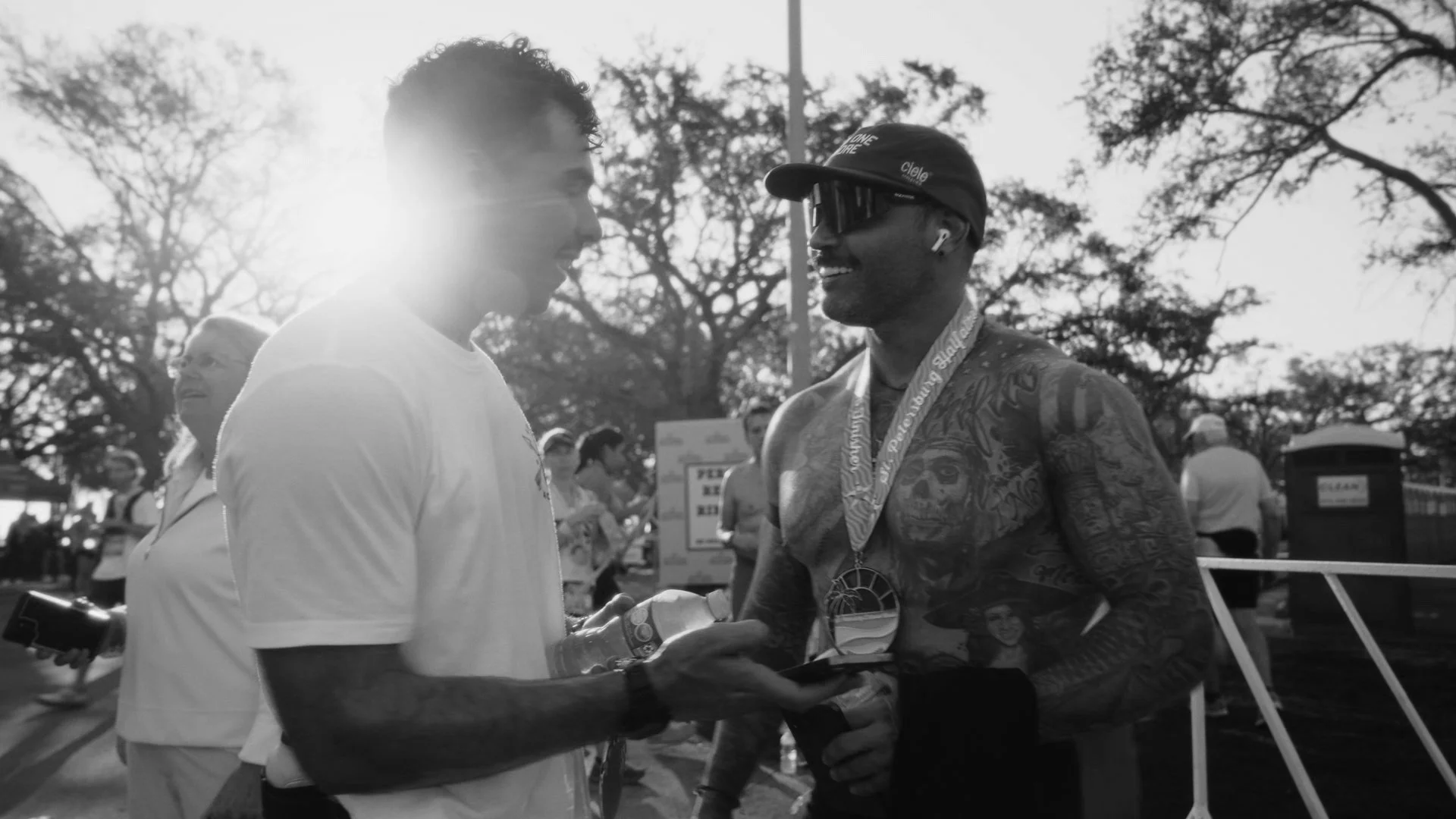Two men conversing outdoors (Anthony Camel & Carlos Contreras), one wearing a cap and sunglasses, the other with curly hair, surrounded by other people and trees in the background, sunlight shining from behind.