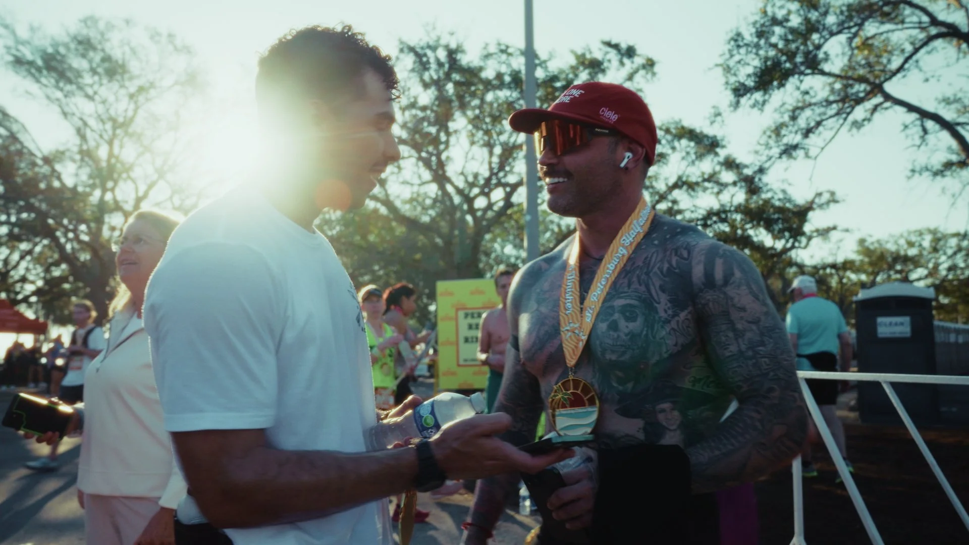 A man with tattoos (Carlos Contreras) is giving a medal to another man (Anthony Camel) at an outdoor event (St. Petersburg Distance Classic) during sunrise.