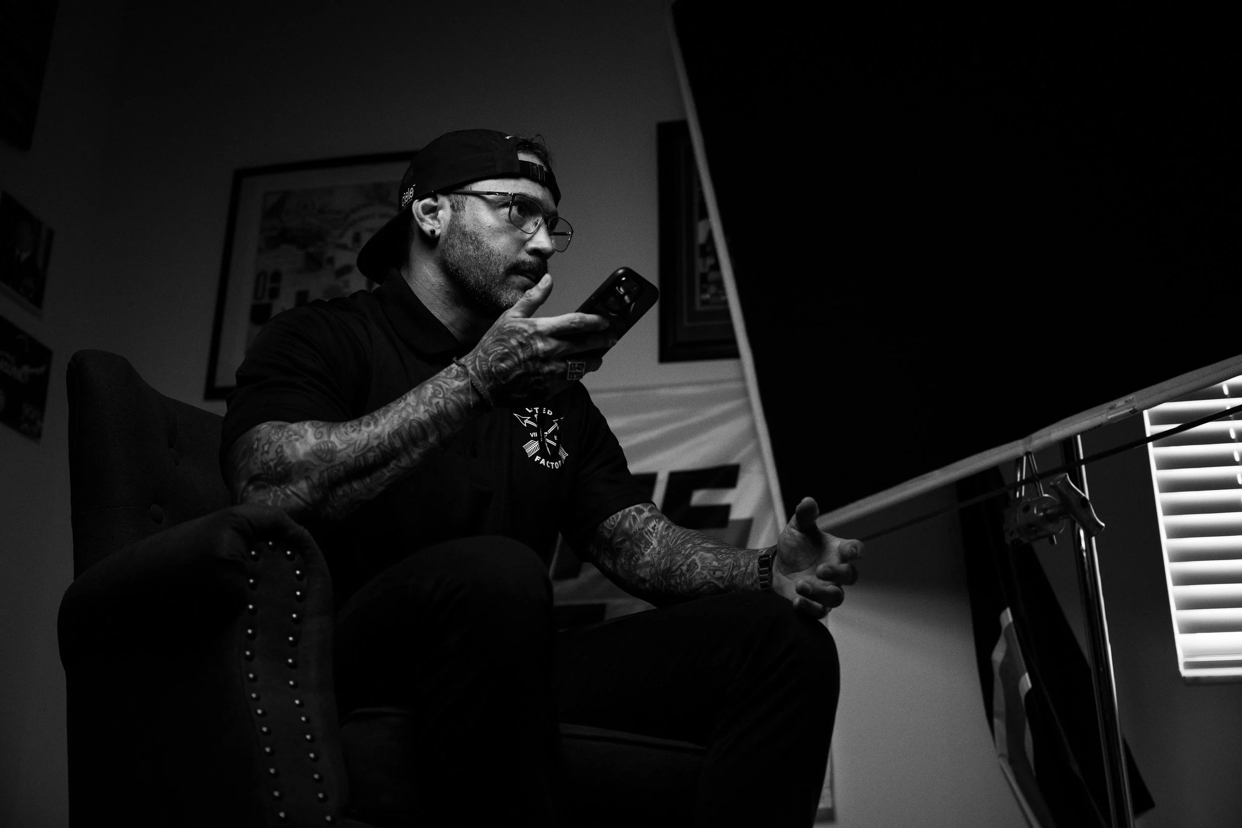 Black and white photo of a man with tattoos (Carlos Contreras), glasses, a backwards cap, and a black polo shirt, sitting on a chair and looking at his phone.