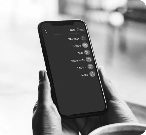 Close-up of a person holding a smartphone displaying a health and fitness app menu with options for workout, cardio, meal, body stats, photos, and sleep in black and white.