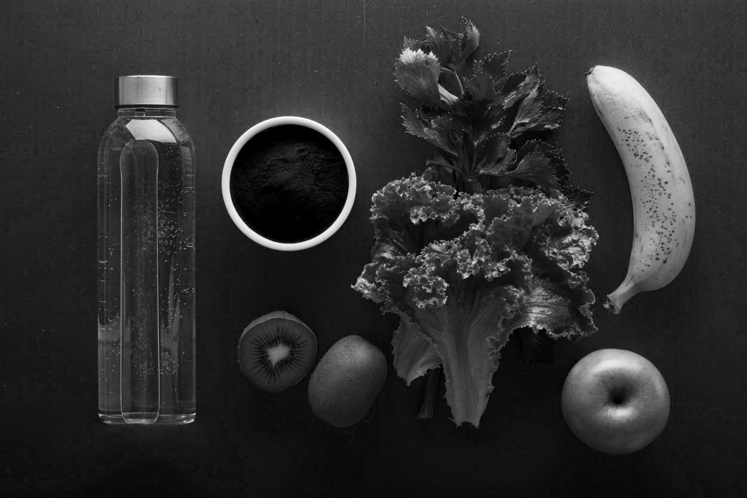 Arrangement of fruits, vegetables, a bottle of water, and a bowl of powder on a dark surface, including an apple, banana, kiwi, leafy greens, and a small bowl with dark powder.