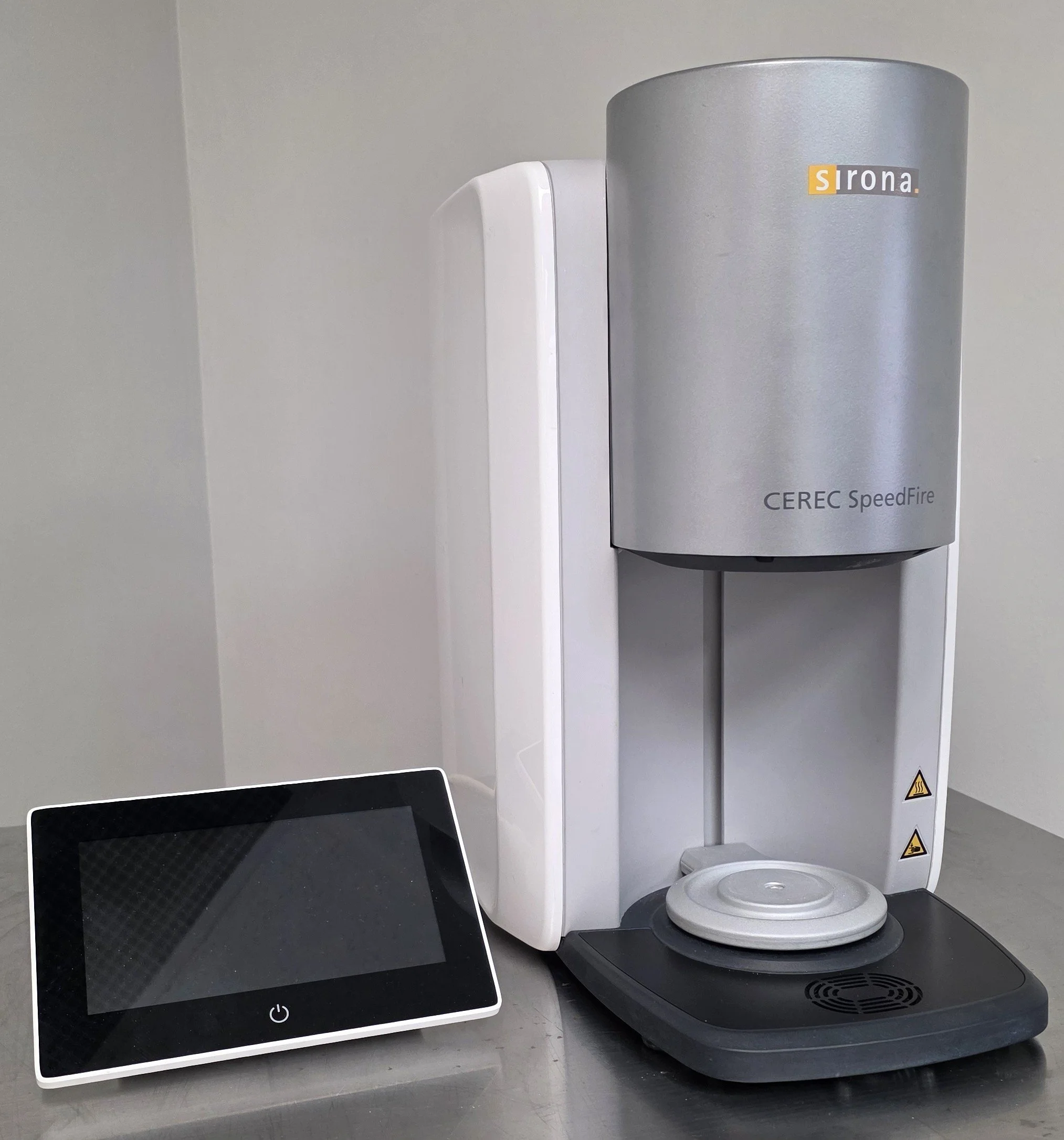 A gray and white 3D printer with the brand name Sirona, model CEREC SpeedFire, is placed on a gray table. To the left of the printer, there is a small black and white touchscreen device laying flat on the table.