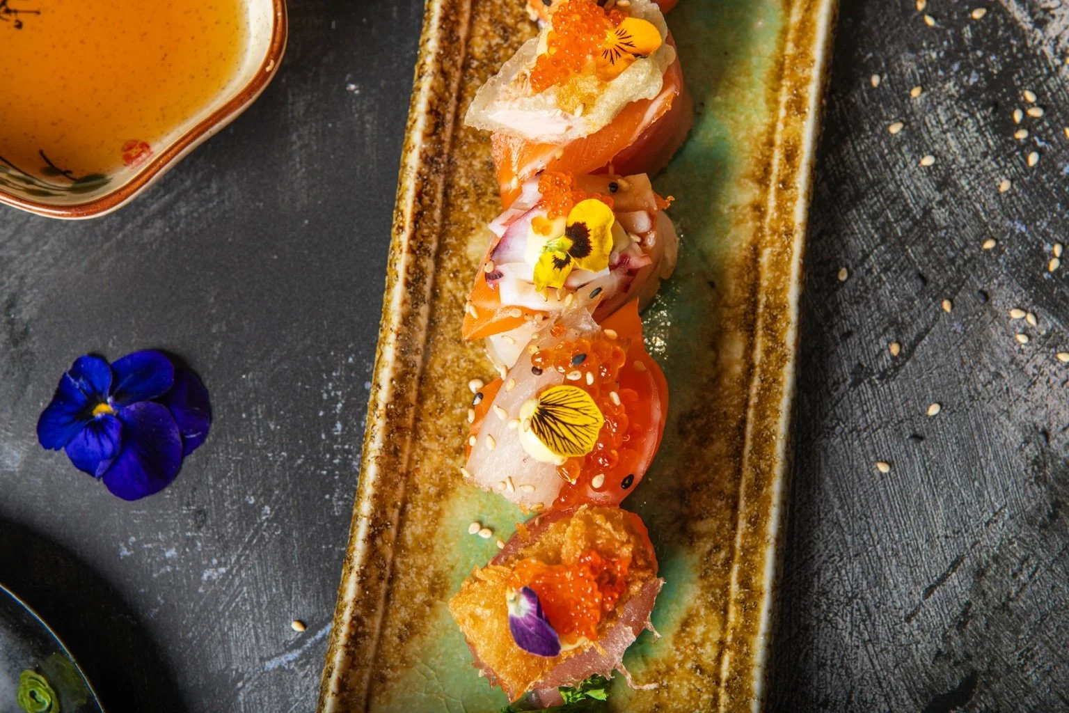Sushi pieces topped with salmon, fish roe and edible flowers on a long ceramic plate.